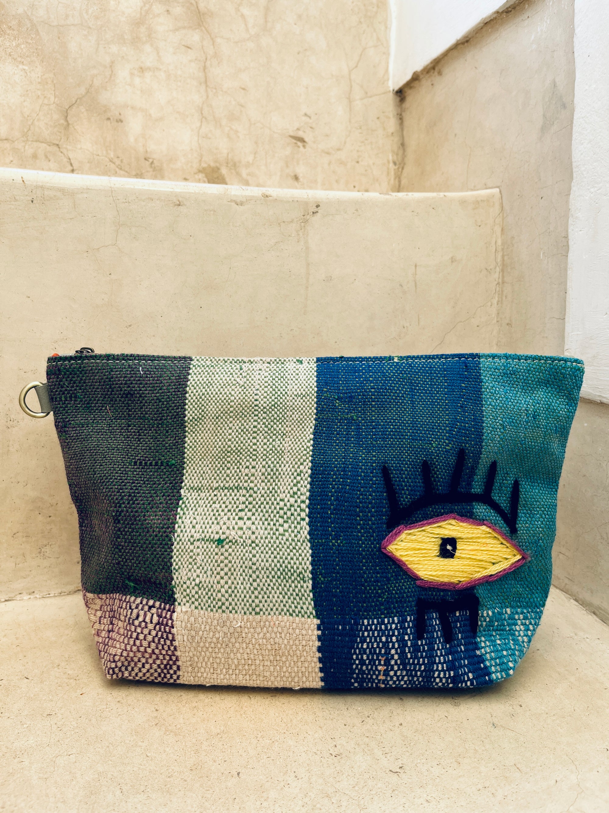 Die LEY! Tasche - Kosmetiktasche zeigt blaue, grüne und beige Streifen sowie ein gelb besticktes Auge mit schwarzen Wimpern auf der Vorderseite und liegt auf beigen Steinstufen.