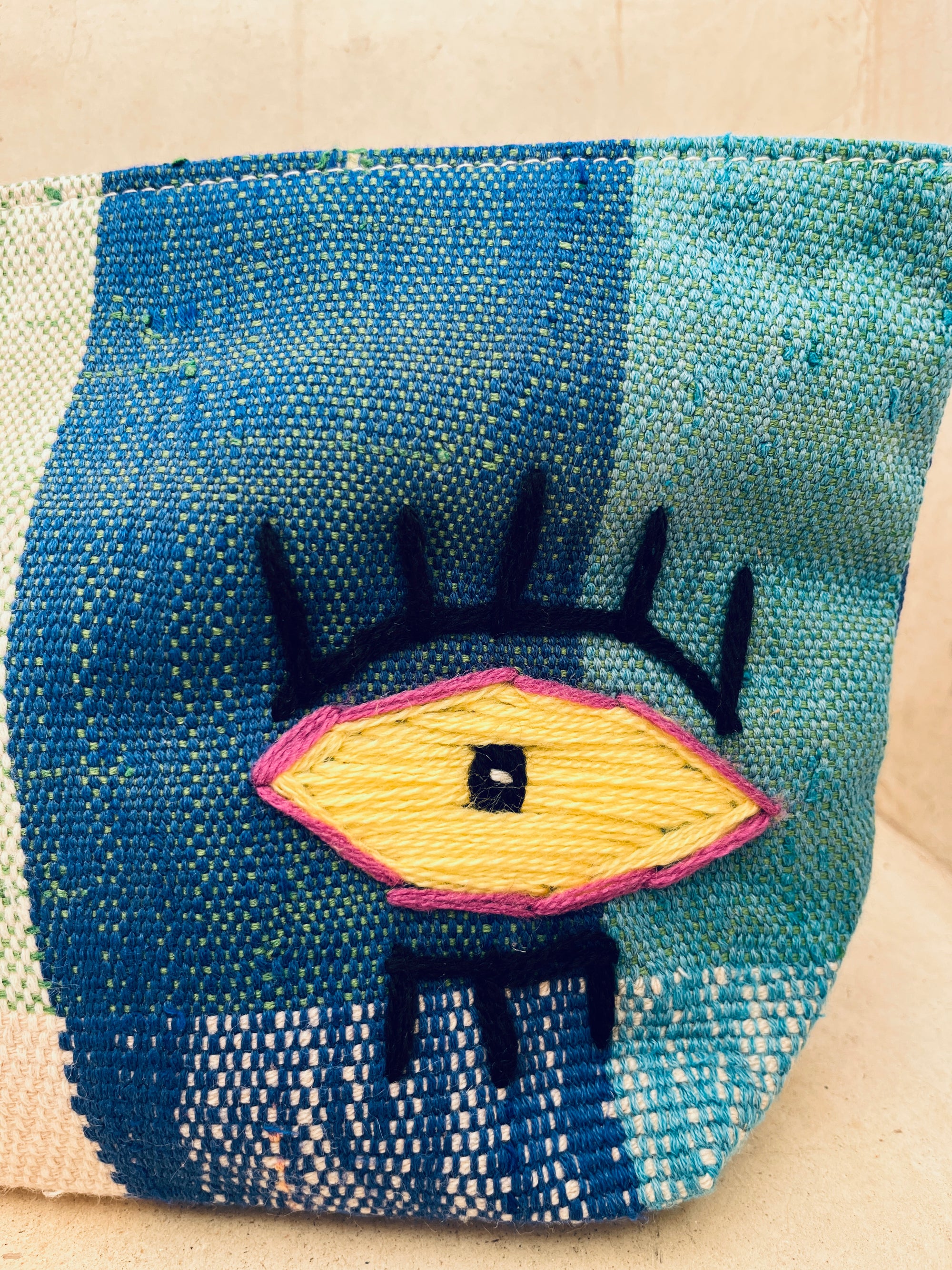 Eine Nahaufnahme der LEY! Tasche - Kosmetiktasche, die ein farbenfrohes gesticktes Auge in Gelb, lila und schwarz umrandet mit schwarzen Wimpern, vor einem Hintergrund aus blauem und grünem gewebtem Stoff zeigt.