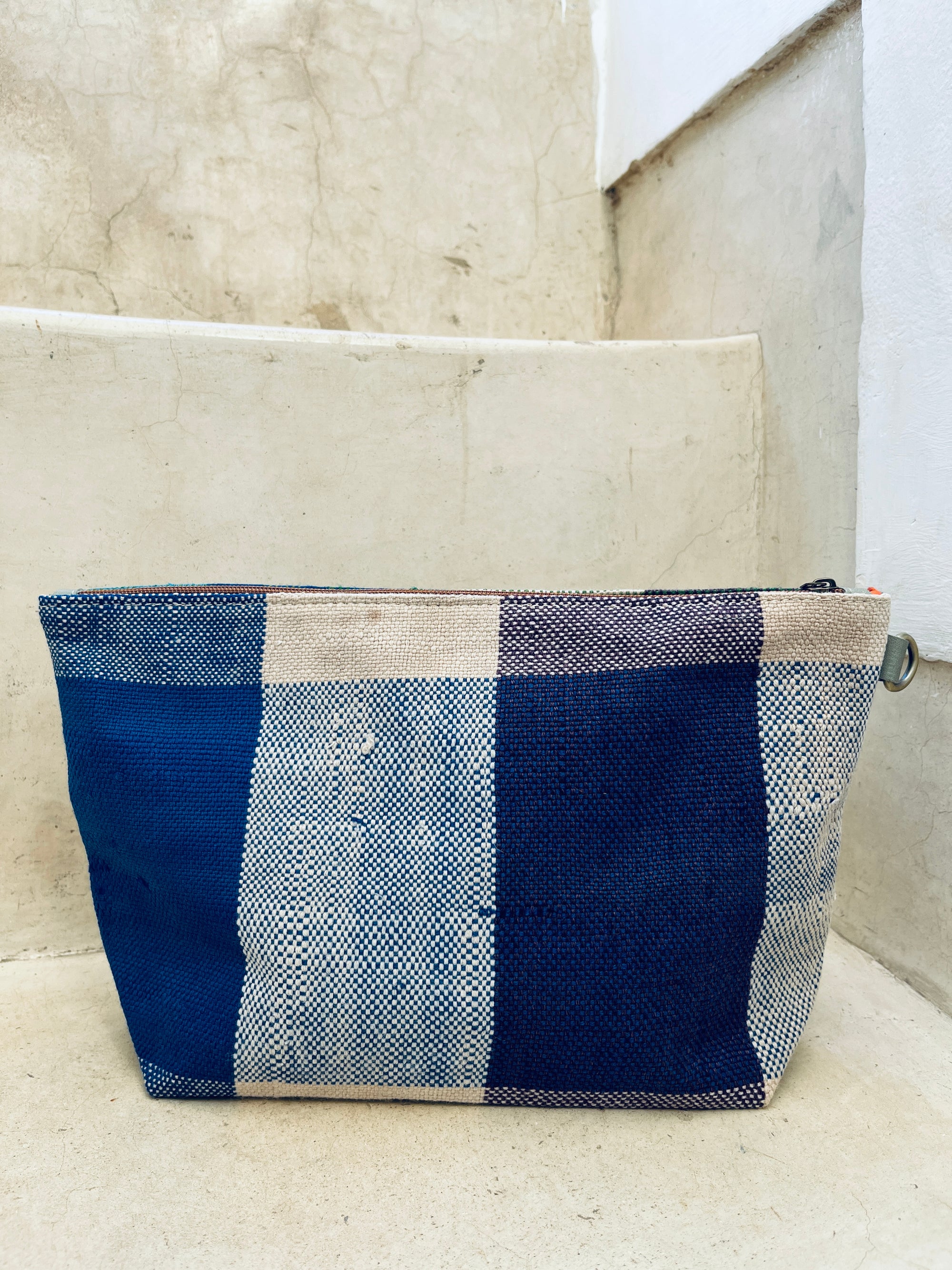 Eine blau-beige karierte Tasche - Kosmetiktasche von LEY! mit Reißverschluss liegt auf hellen Betonstufen vor einer strukturierten Wand.
