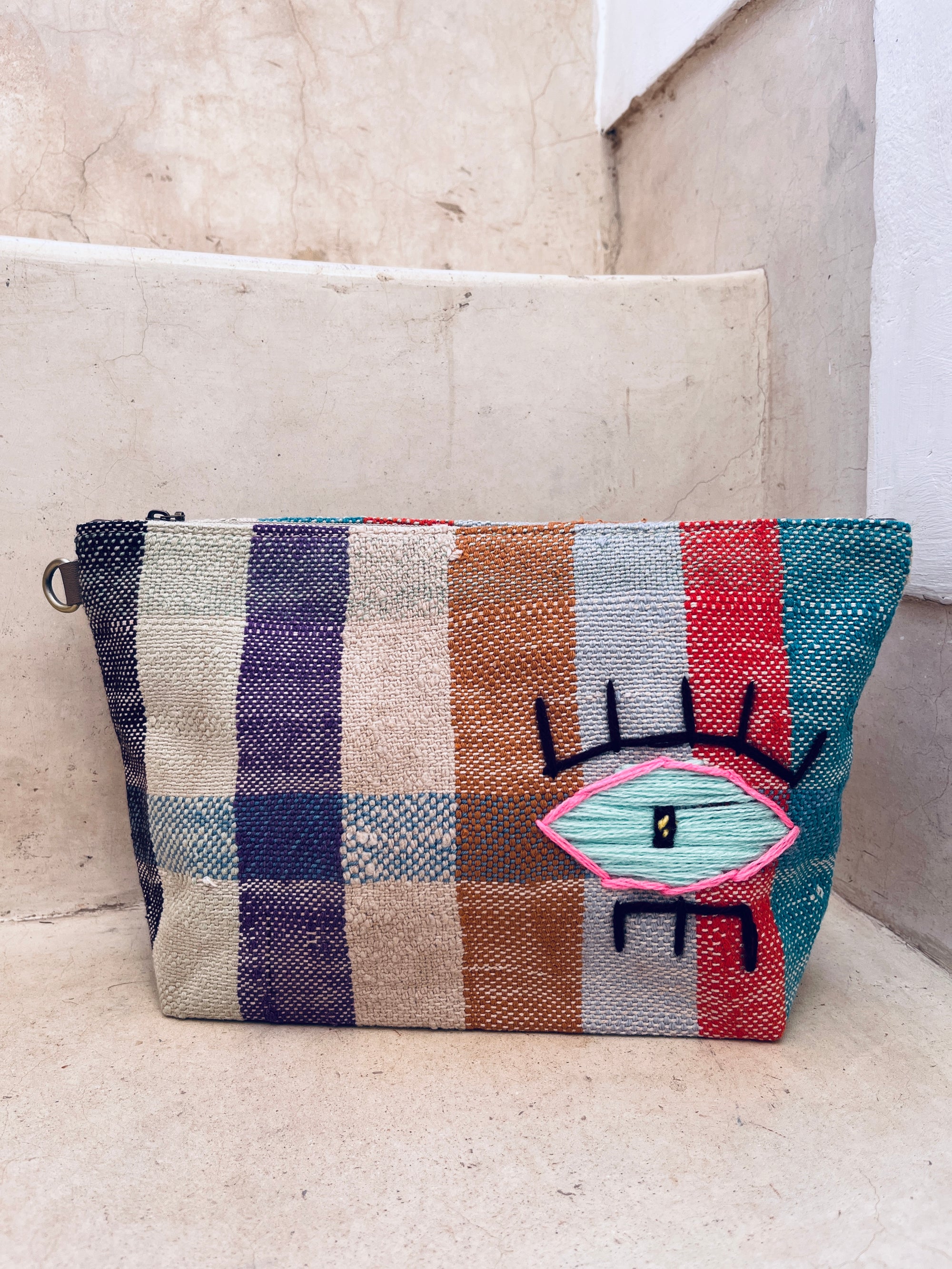 Die handgewebte Kosmetiktasche von LEY! besticht durch beige, blaue, orange und rote Streifen sowie ein gesticktes Auge in Türkis, Weiß und Pink. Das stilvolle Accessoire aus Marokko ist auf Steinstufen platziert.