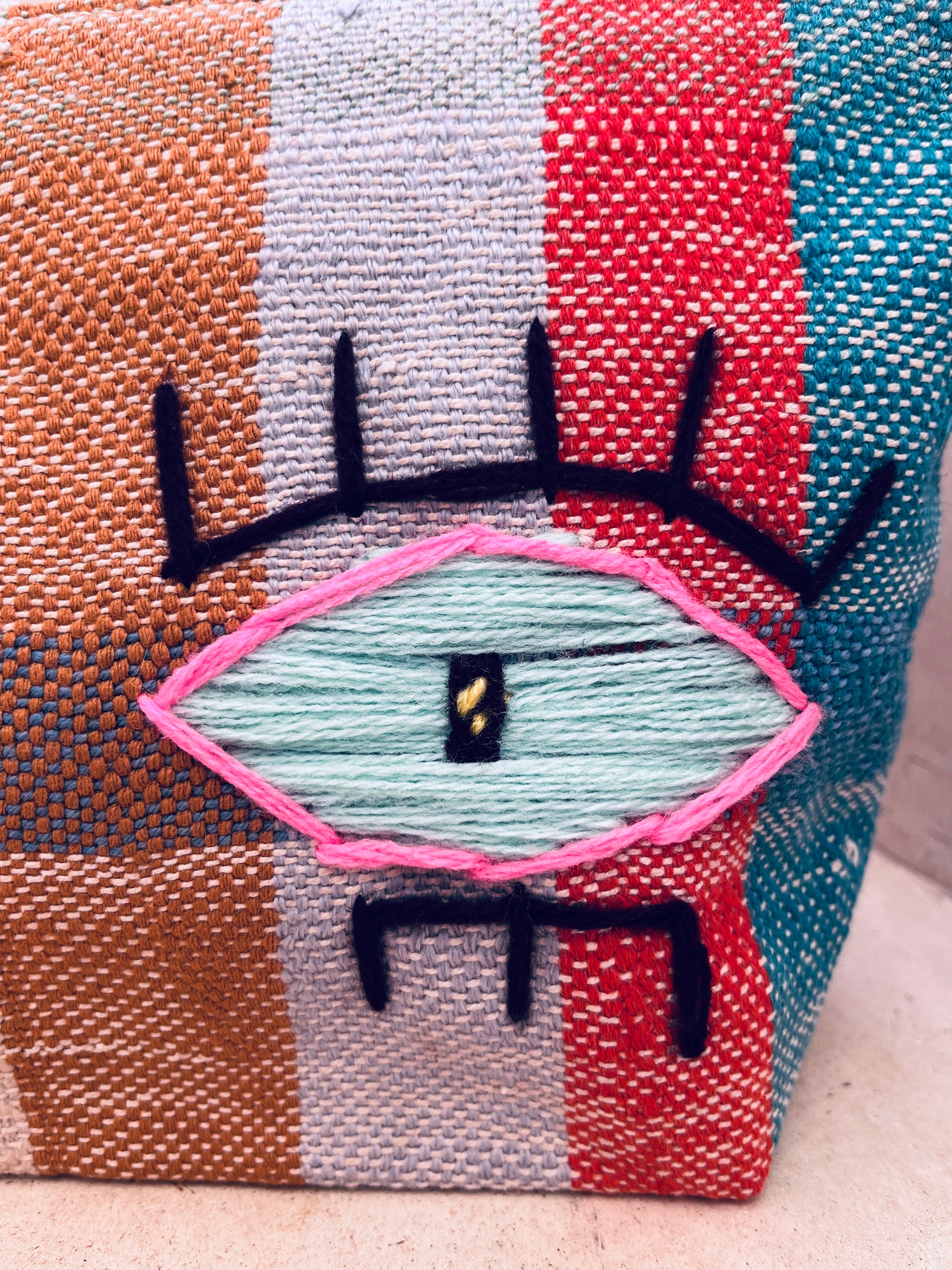 Die LEY! Tasche - Kosmetiktasche besticht durch vertikale Streifen in Orange, Beige, Blau und Türkis sowie ein gesticktes, stilisiertes Auge mit pinker Umrandung, schwarzen Wimpern und gelber Pupille.