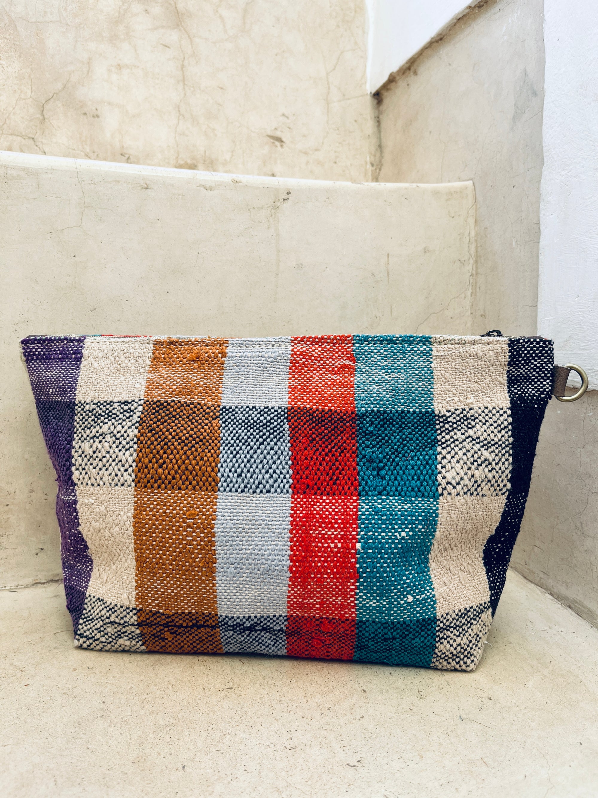 Die LEY! Tasche - Kosmetiktasche zeigt vertikale Streifen in Lila, Orange, Grau, Rot, Petrol und Beige und ruht auf hellen Steinstufen vor einer passenden Wand.