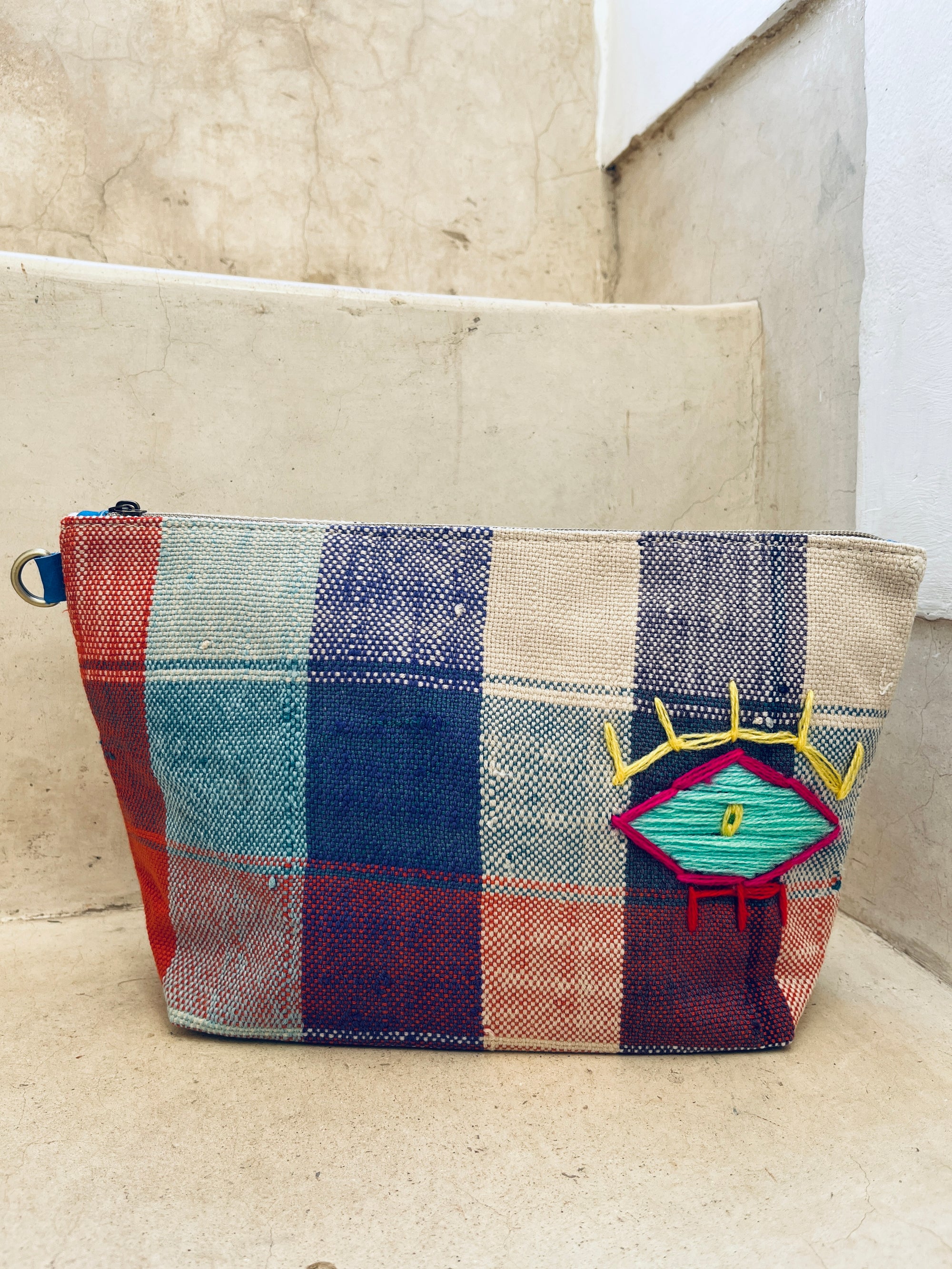 Die LEY! Tasche - Kosmetiktasche ist eine bunte, handgefertigte Kosmetiktasche mit Reißverschluss, blau-roten Streifen und einem gestickten Augendesign mit gelben Wimpern und türkisem Zentrum auf der Vorderseite.