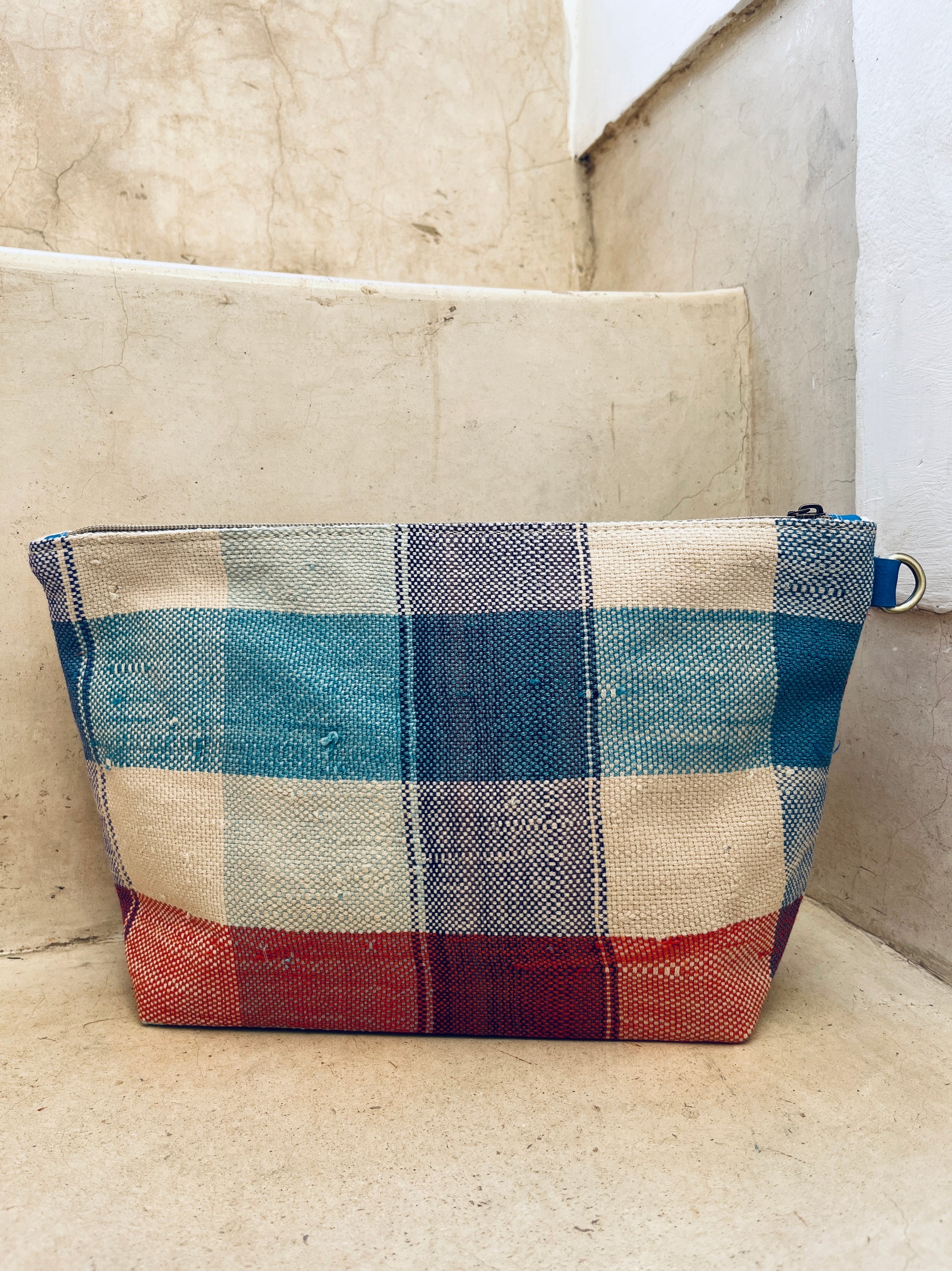 Die handgefertigte Kosmetiktasche "Tasche - Kosmetiktasche" von LEY! zeigt ein kariertes Muster in Blau, Beige und Rot. Sie liegt auf einer beigen Betonstufe an einer weißen Wand, inspiriert vom traditionellen marokkanischen Design.