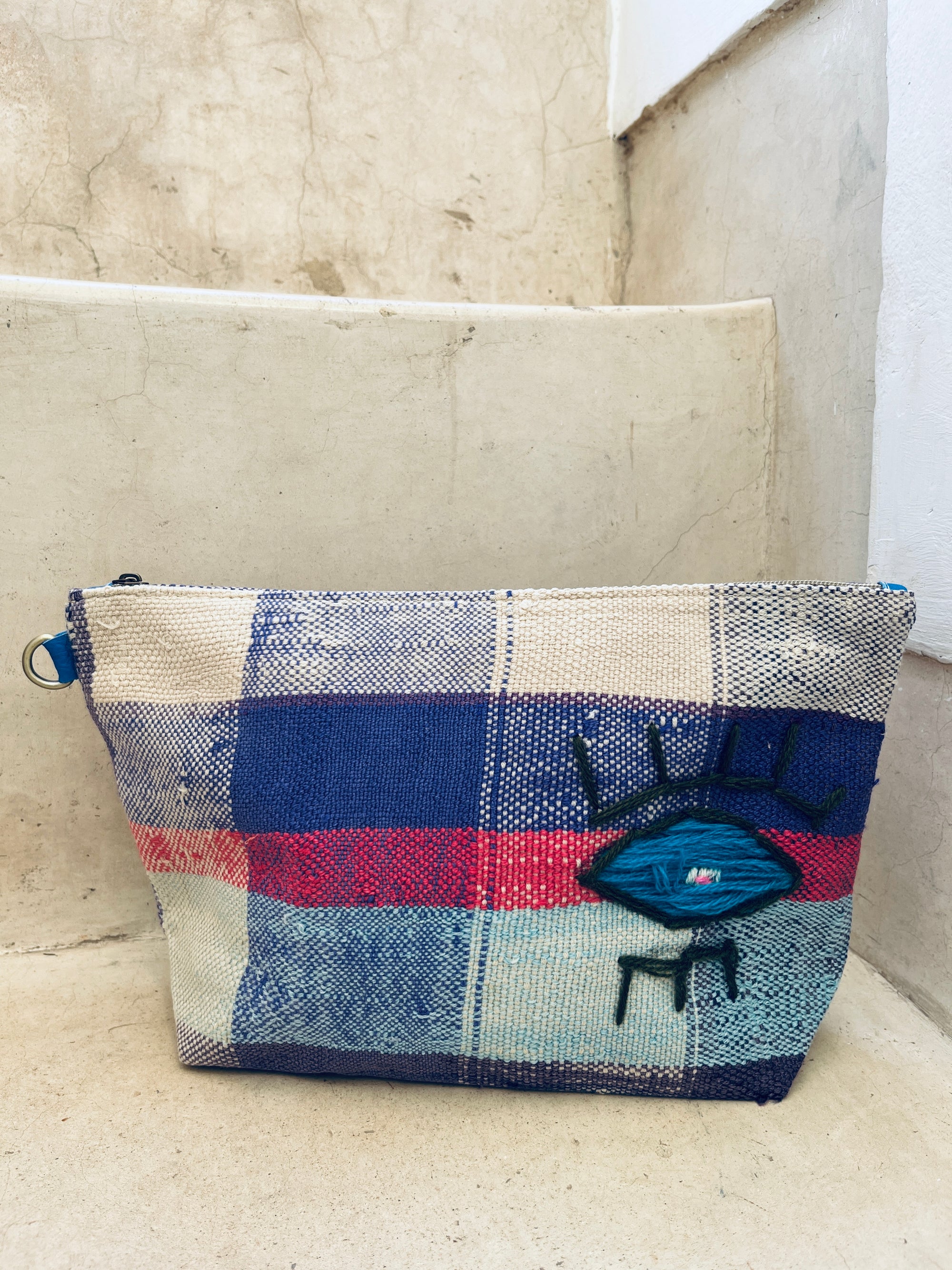 Die LEY! Kosmetiktasche zeigt auffälliges Auge-Motiv mit blauen Wimpern und schwarzen Beinen auf blau-rotem und cremefarbenem Plaidmuster. Handgefertigt in Marokko, fotografiert auf Steinstufen.