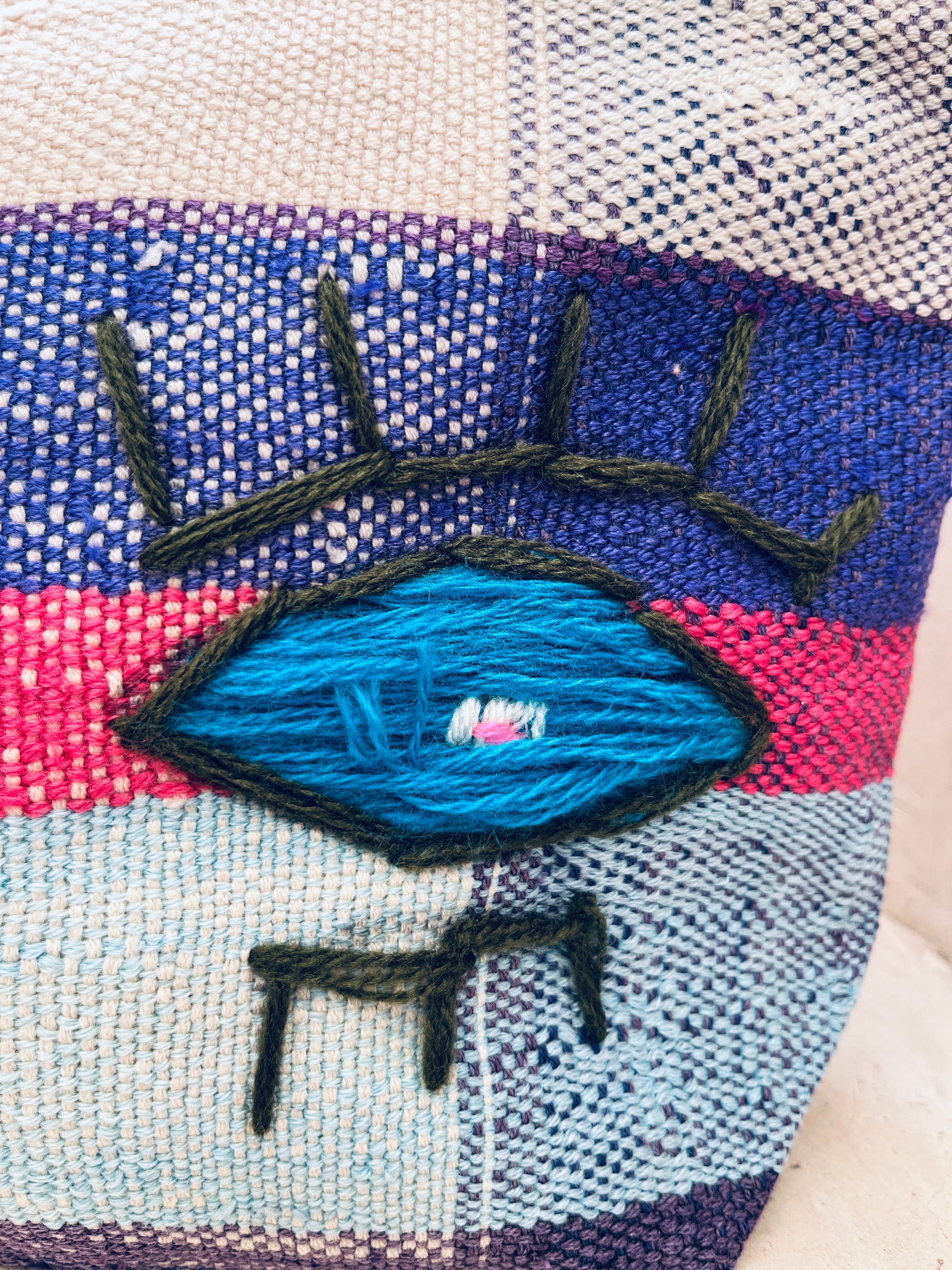 Eine Nahaufnahme der Tasche - Kosmetiktasche von LEY! zeigt ein gesticktes, stilisiertes Auge in Schwarz und Blau auf pink-blau-creme gestreiftem, handgewebtem Stoff.