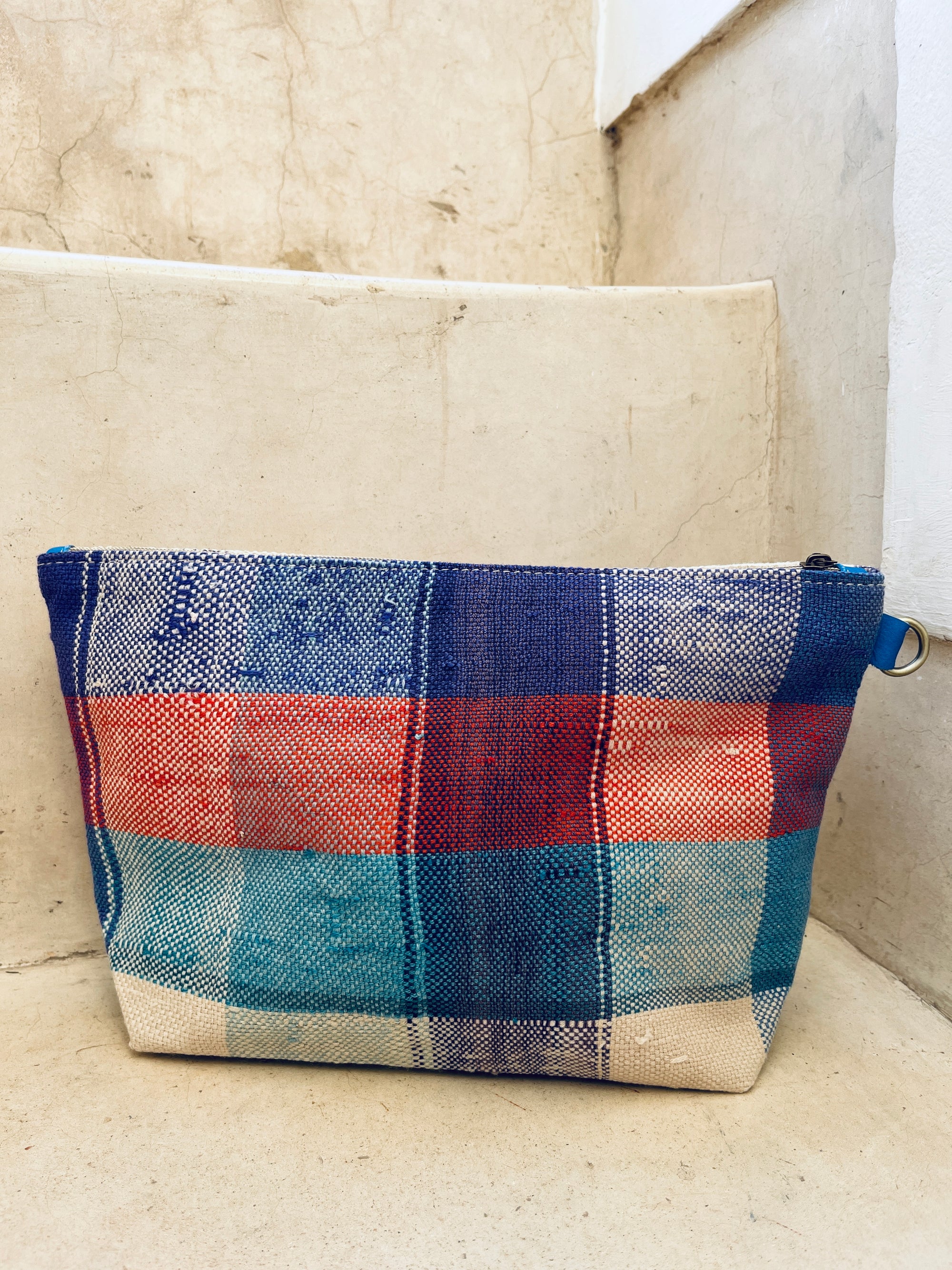 Die LEY! Tasche - Kosmetiktasche mit blau-rotem und weißem Plaidmuster liegt auf beigen Betonstufen.