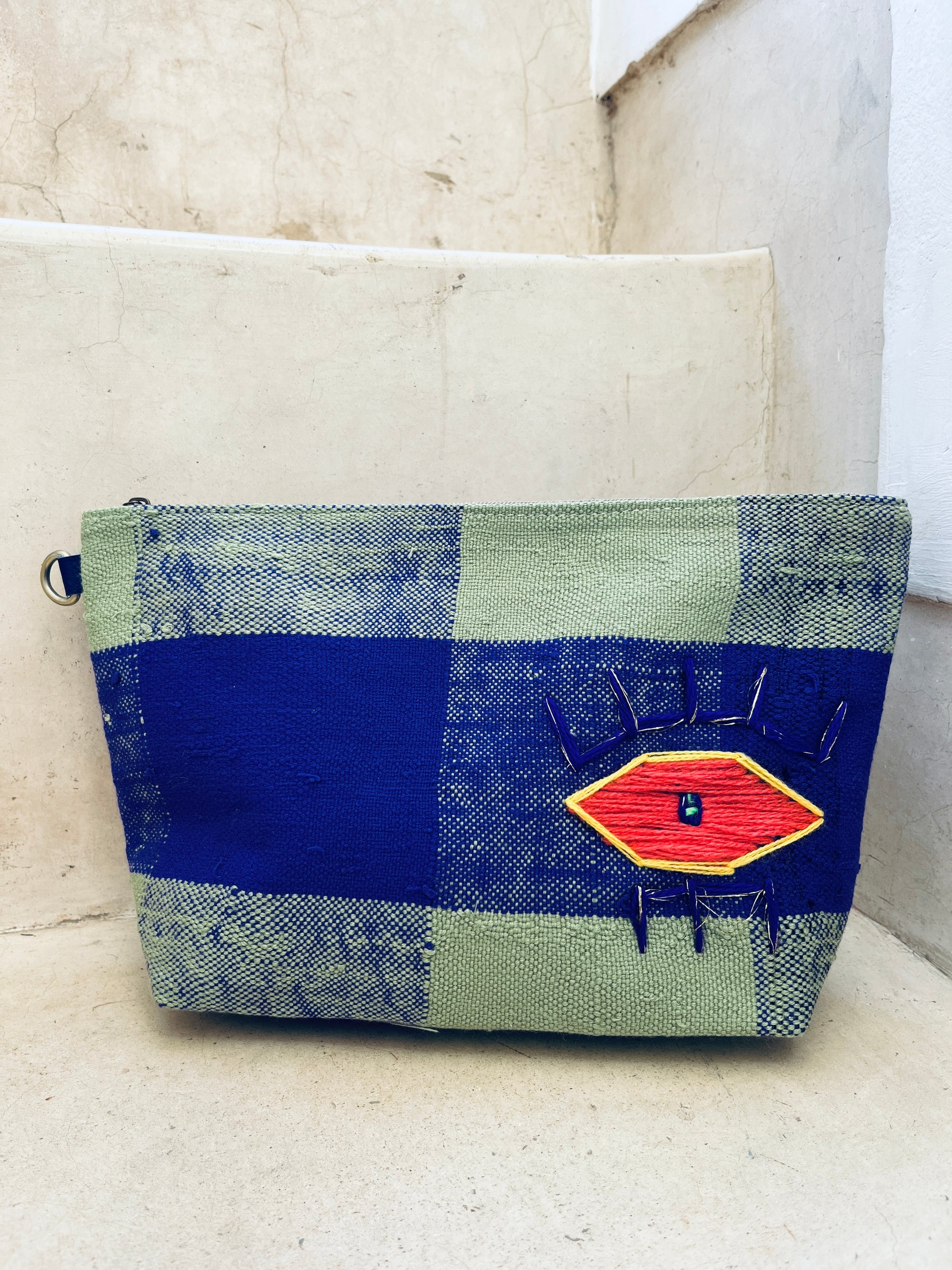 Die Kosmetiktasche "Tasche - Kosmetiktasche" von LEY! zeigt ein auffälliges, rot-gelb gesticktes Auge-Motiv mit schwarzen Nähten auf grün-blau kariertem Stoff und liegt auf Steinstufen.