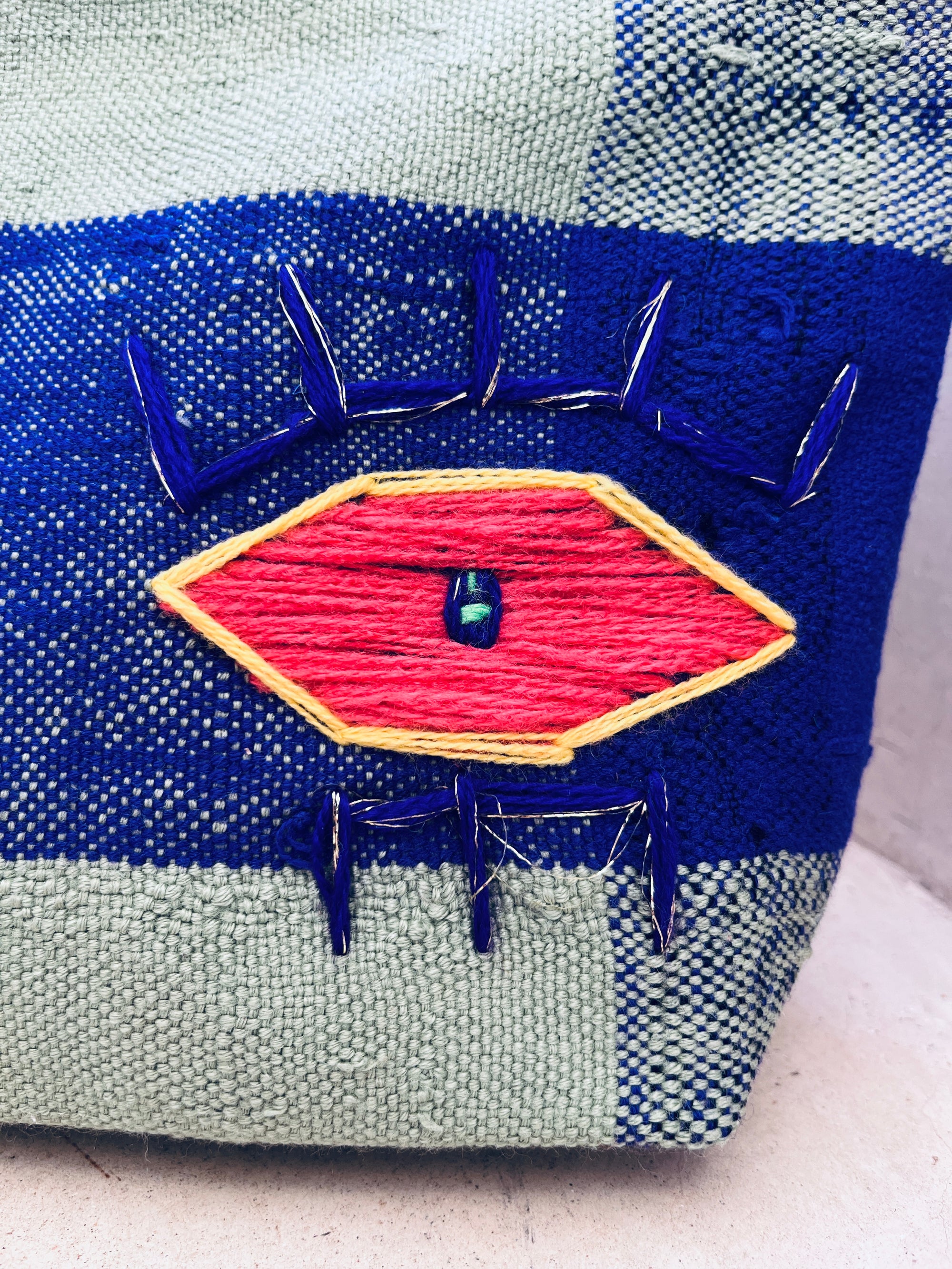 Eine Nahaufnahme der LEY! Tasche - Kosmetiktasche, handgewebt mit einem hellblauen und hellgrünen Karomuster und einem großen bunten Augenmotiv, gestickt mit rotem, gelbem und blauem Garn und einer kleinen zentralen Perle.