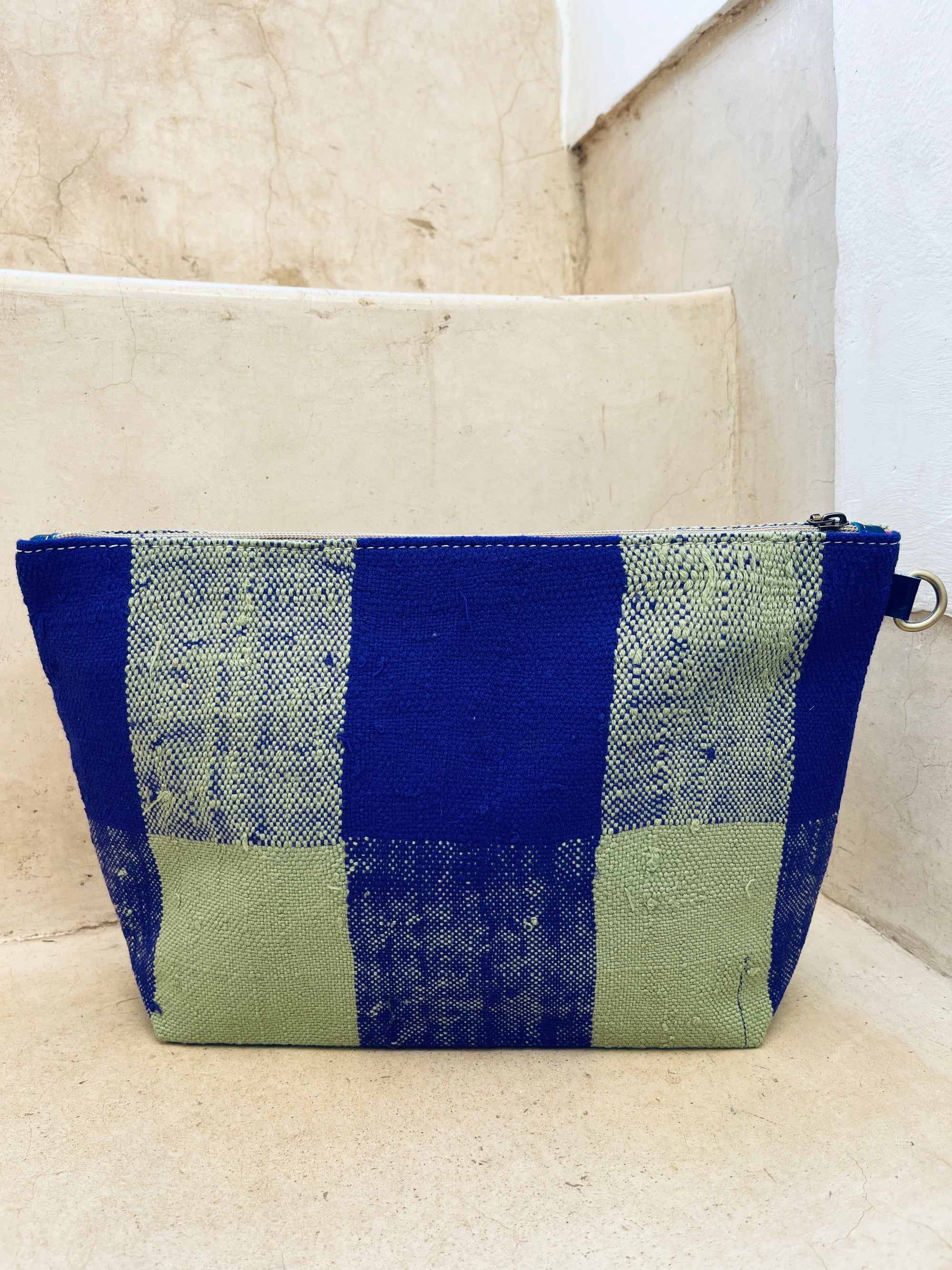 Die rechteckige LEY! Tasche - Kosmetiktasche besticht durch ihr blau-grün kariertes, handgewebtes Muster im marokkanischen Stil und hat oben einen Reißverschluss. Sie liegt auf beigen Steinstufen.