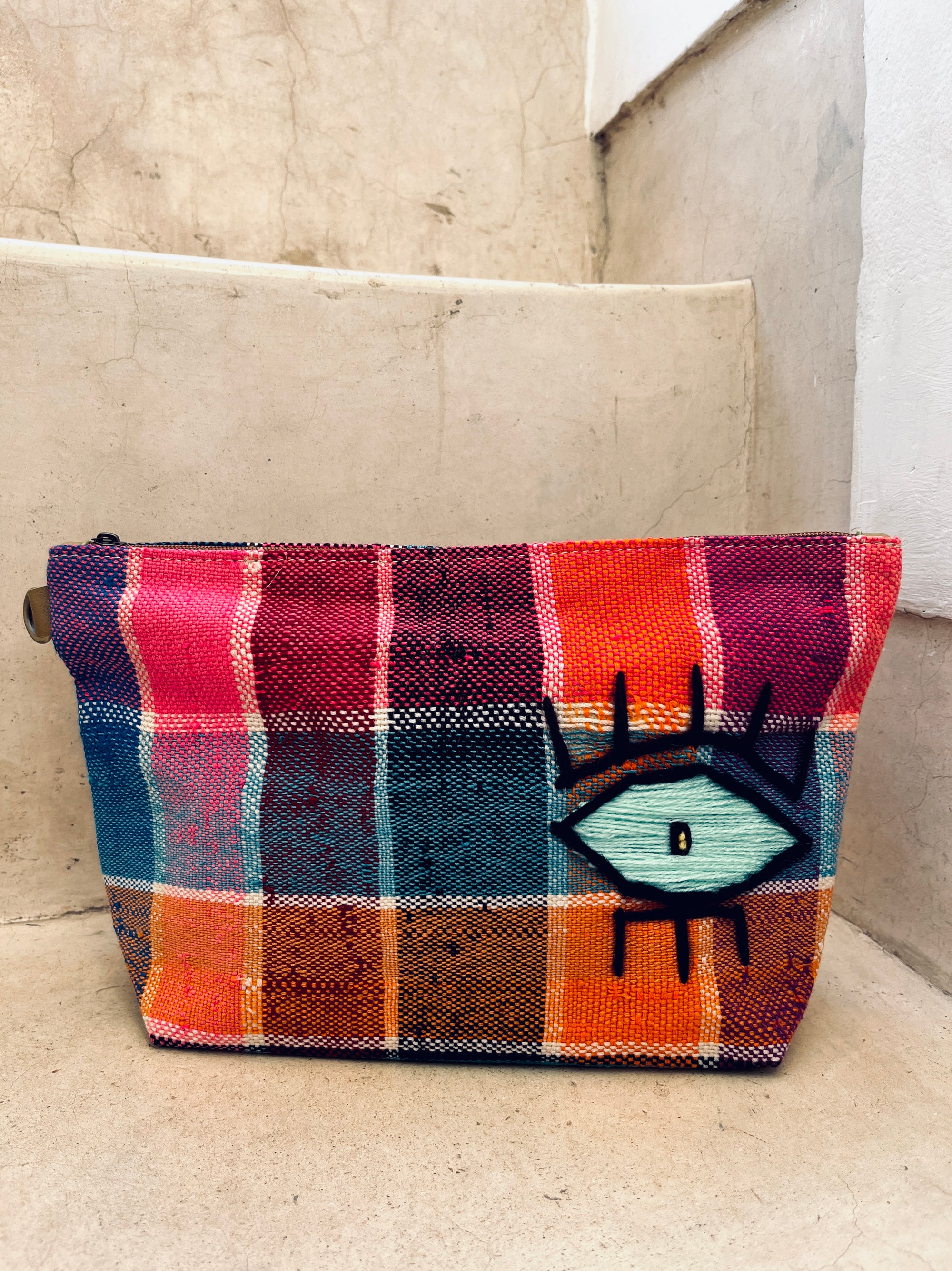 Die Tasche - Kosmetiktasche von LEY! ist eine handgewebte Kosmetiktasche aus Marokko mit Tartanmuster in Rot, Orange, Blau und Lila sowie auffälligem Auge-Motiv in Schwarz, Weiß und Türkis.