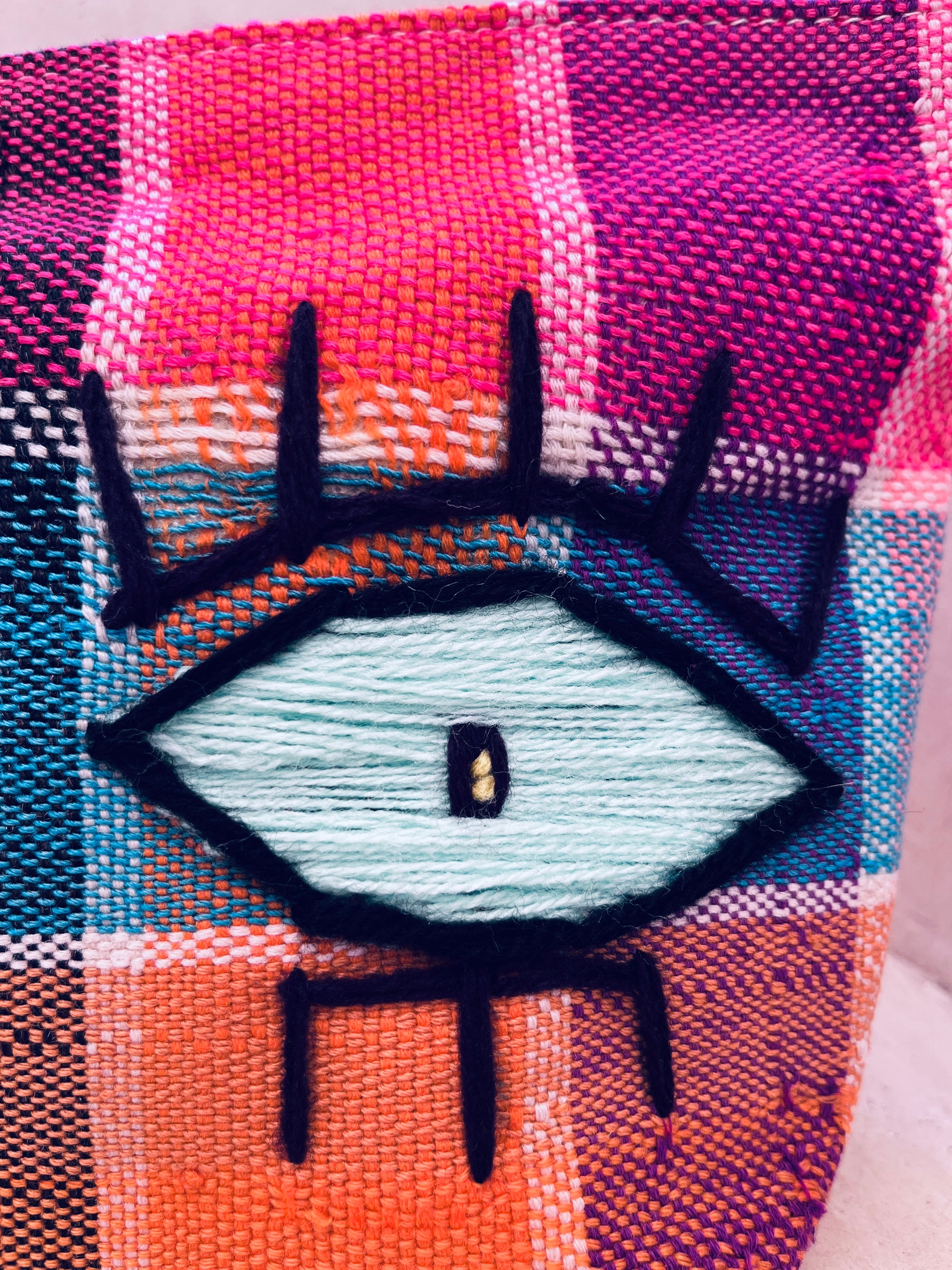 Nahaufnahme der LEY! Tasche - Kosmetiktasche: handgewebt in Marokko, mit großem stilisiertem Auge aus schwarzem Garn auf kariertem Muster in Pink, Orange, Blau und Weiß.