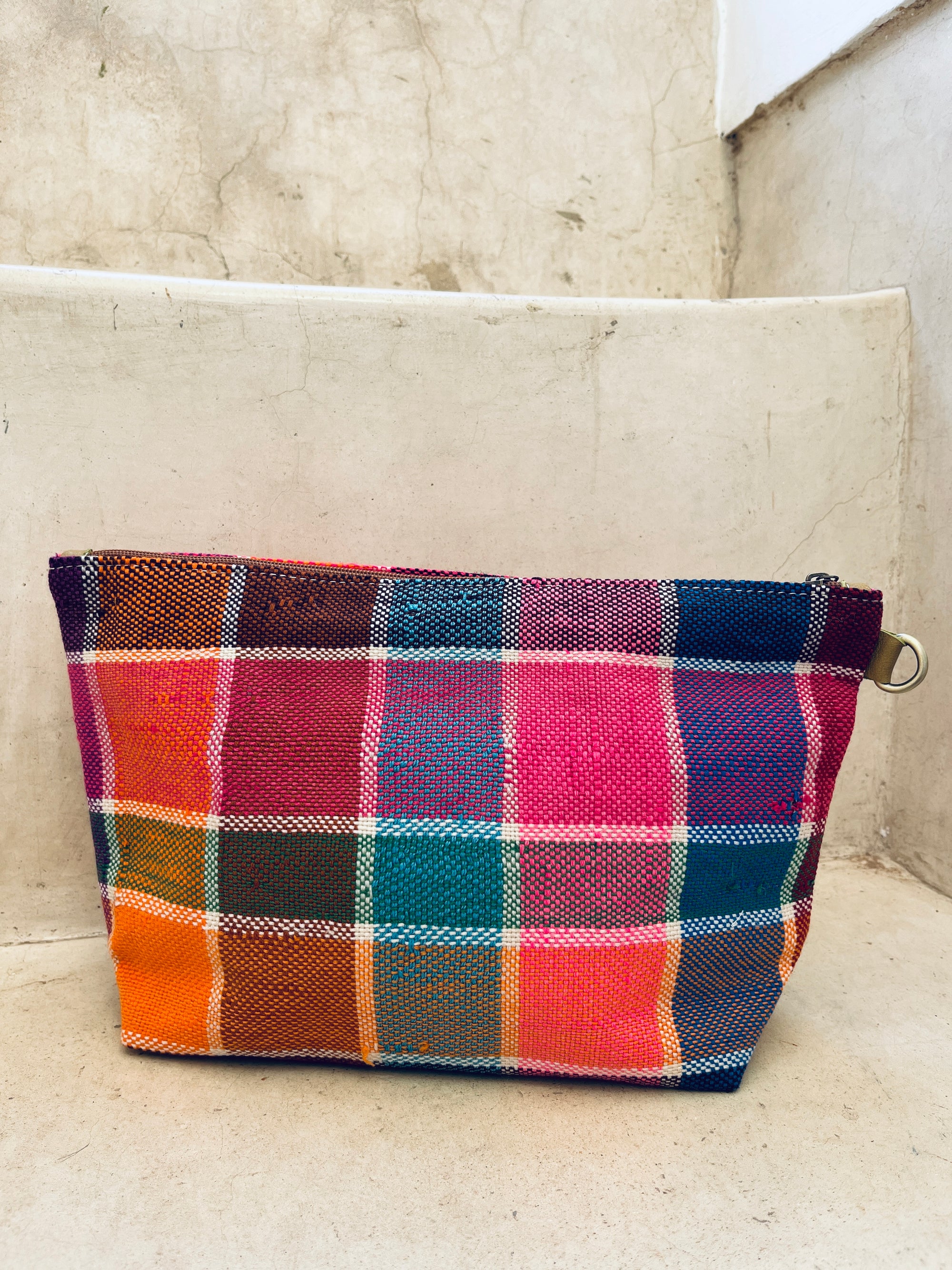 Die LEY! Tasche - Kosmetiktasche ist handgewebt in Marokko, zeigt Plaidmuster in Orange, Pink, Rot, Blau und Grün und besitzt einen Metallring. Sie liegt auf hellen Betontreppen.
