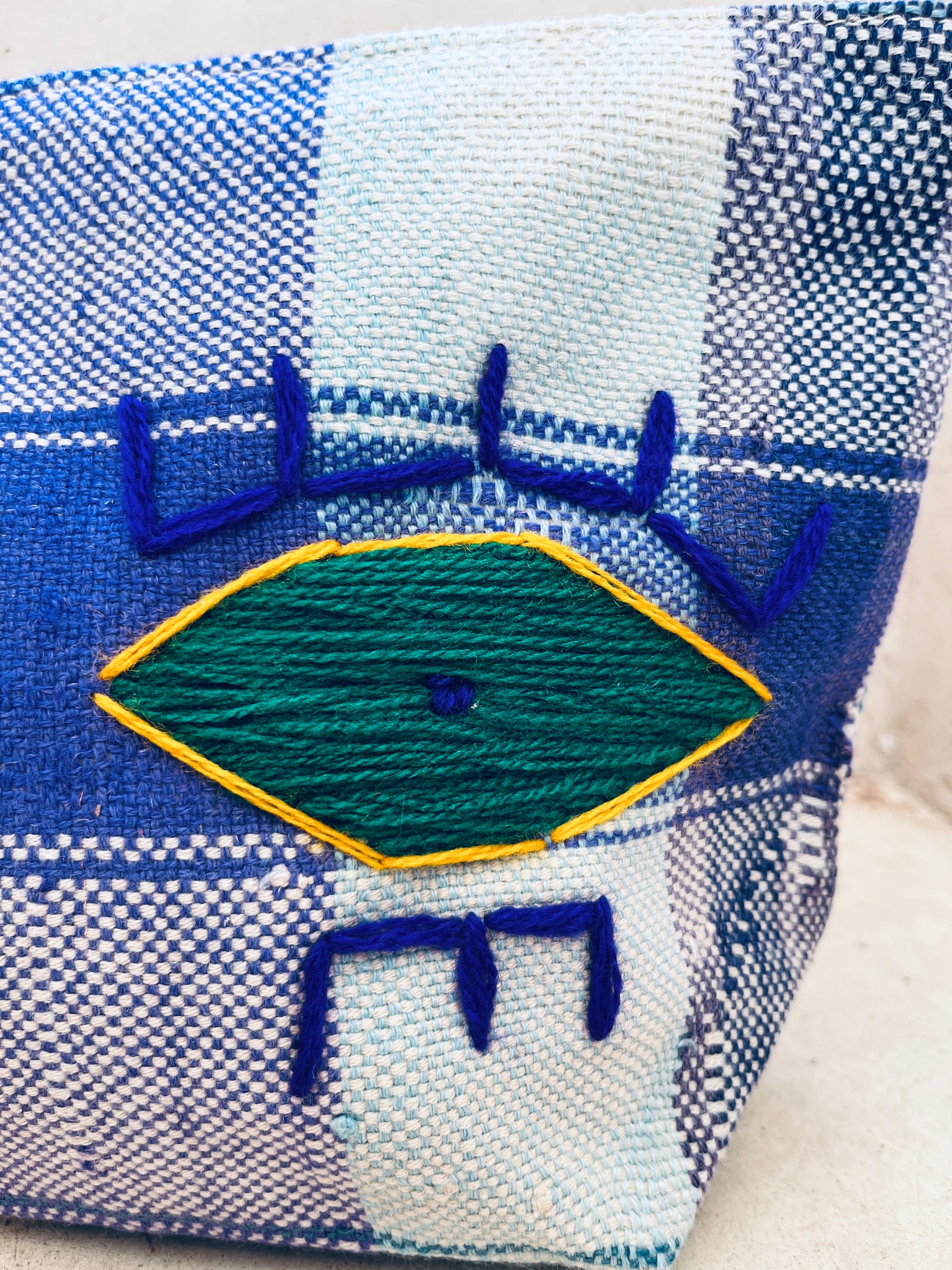 Eine Nahaufnahme eines handgewebten blau-weiß karierten Stoffes mit einer von den Augen inspirierten geometrischen Stickerei - einer grünen, gelb umrandeten Raute mit blauen Garndetails - ziert die LEY! Tasche - Kosmetiktasche.