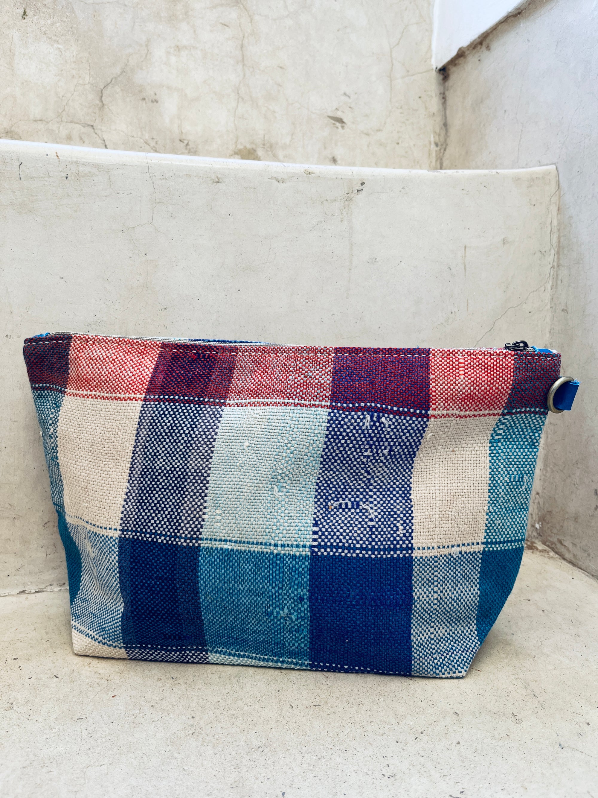 Eine rechteckige, handgefertigte Tasche - Kosmetiktasche von LEY! mit Reißverschluss und rot-weiß-blauem Karomuster aus geflochtenem Kunststoff liegt auf einer hellen Betonstufe.
