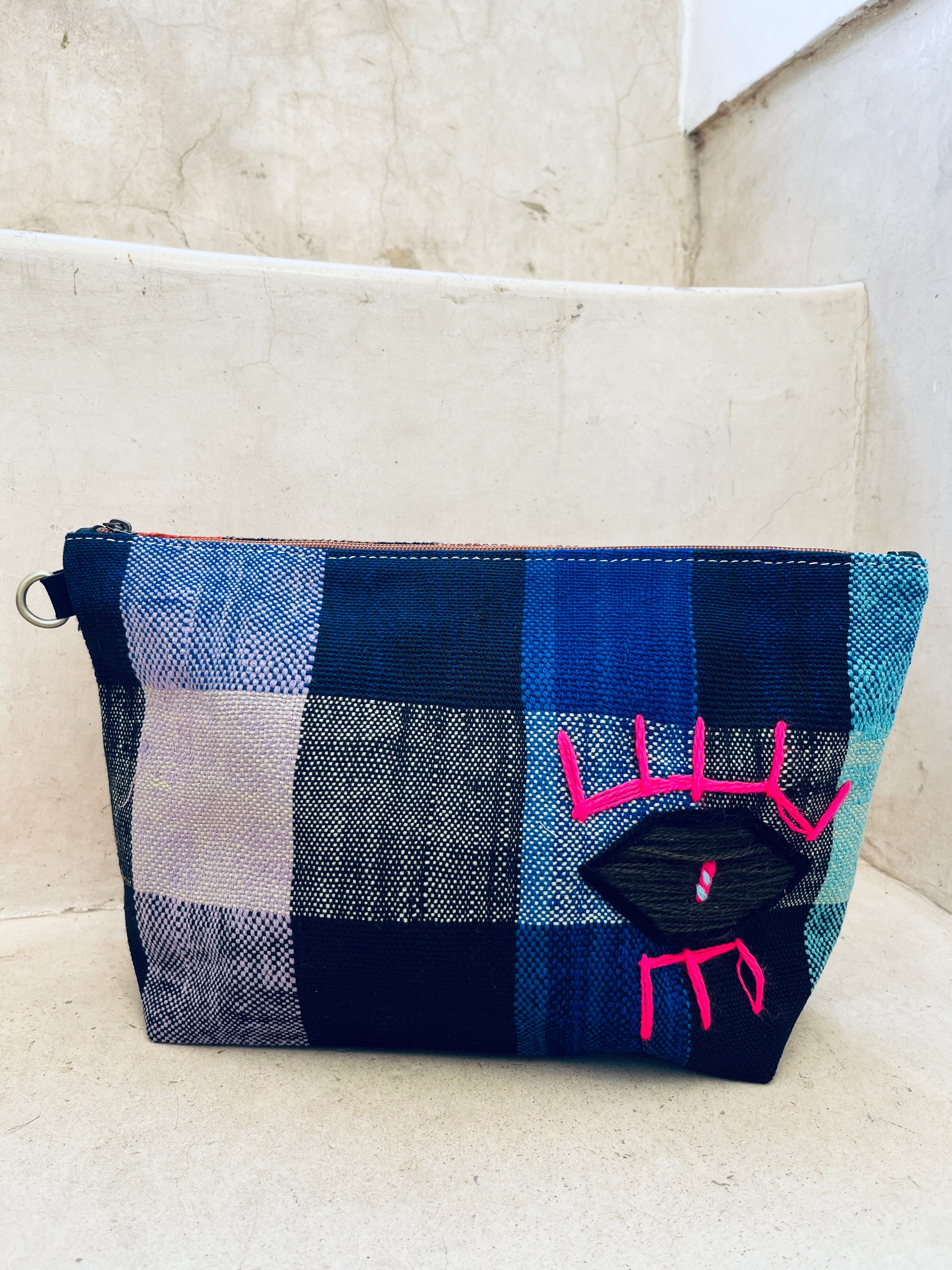 Die LEY! Tasche - Kosmetiktasche ist eine blau-schwarz karierte Reißverschlusstasche auf einer weißen Steintreppe mit einem handgestickten schwarzen Auge mit rosa Wimpern auf der Vorderseite.