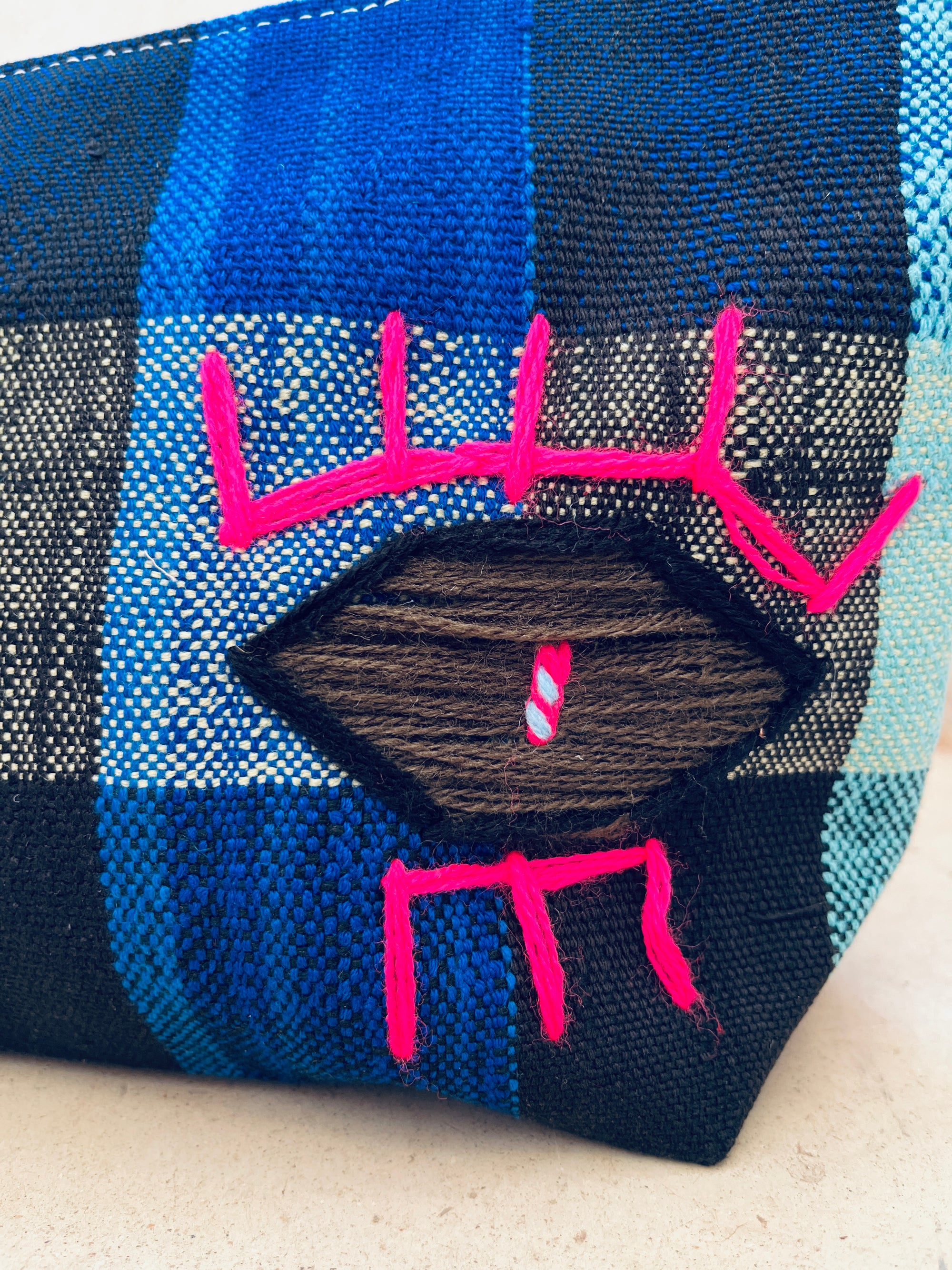 Eine Nahaufnahme der LEY! Tasche - Kosmetiktasche zeigt strukturierten blau-schwarz karierten Stoff, handgewebt, mit einem schwarzen Rautenaufnäher und leuchtend rosa abstrakten Stickereien, die dieses Stück von Marokko wirklich einzigartig machen.