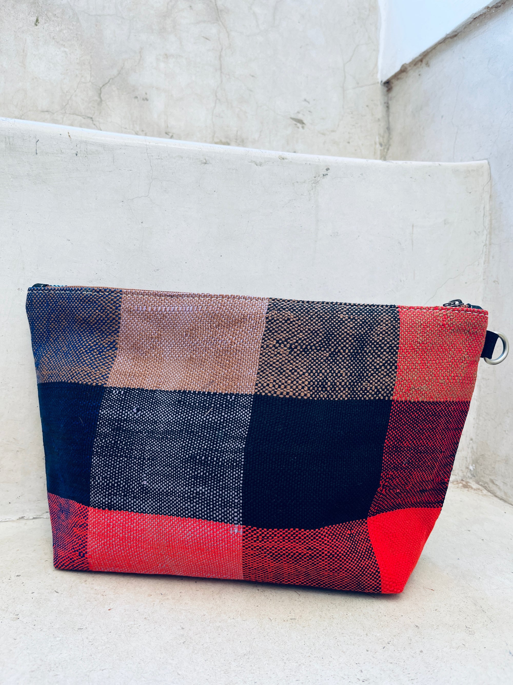 Die handgefertigte rechteckige Tasche - Kosmetiktasche von LEY! mit rotem, schwarzem, braunem und beigem Karomuster liegt auf hellen Betontreppen.
