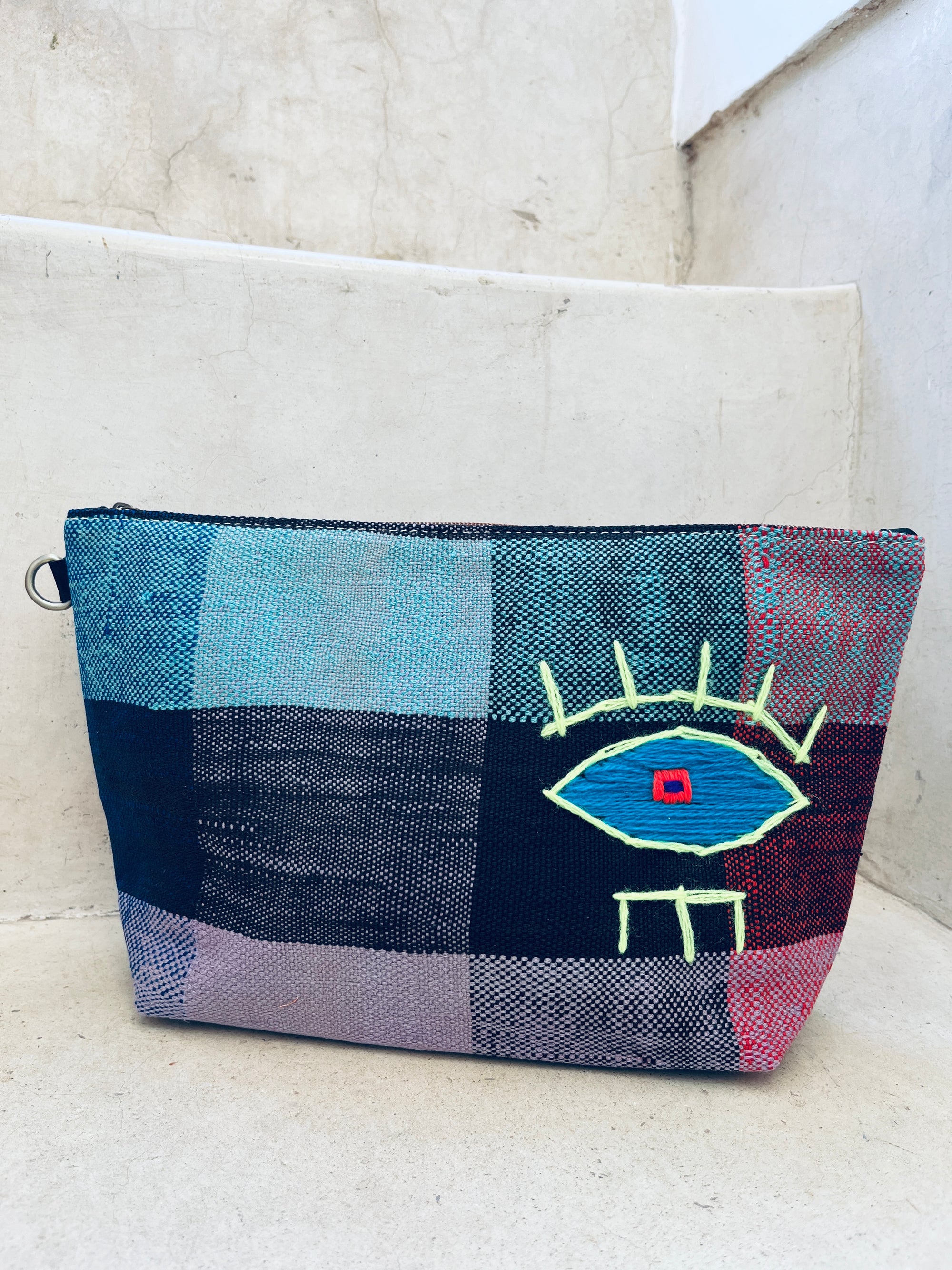 Die handgewebte Tasche - Kosmetiktasche von LEY! aus Marokko zeigt ein abstrakt gesticktes Auge in Gelb, Blau und Rot auf blau-schwarz-pink-rotem Karo und liegt auf weißen Steinstufen.