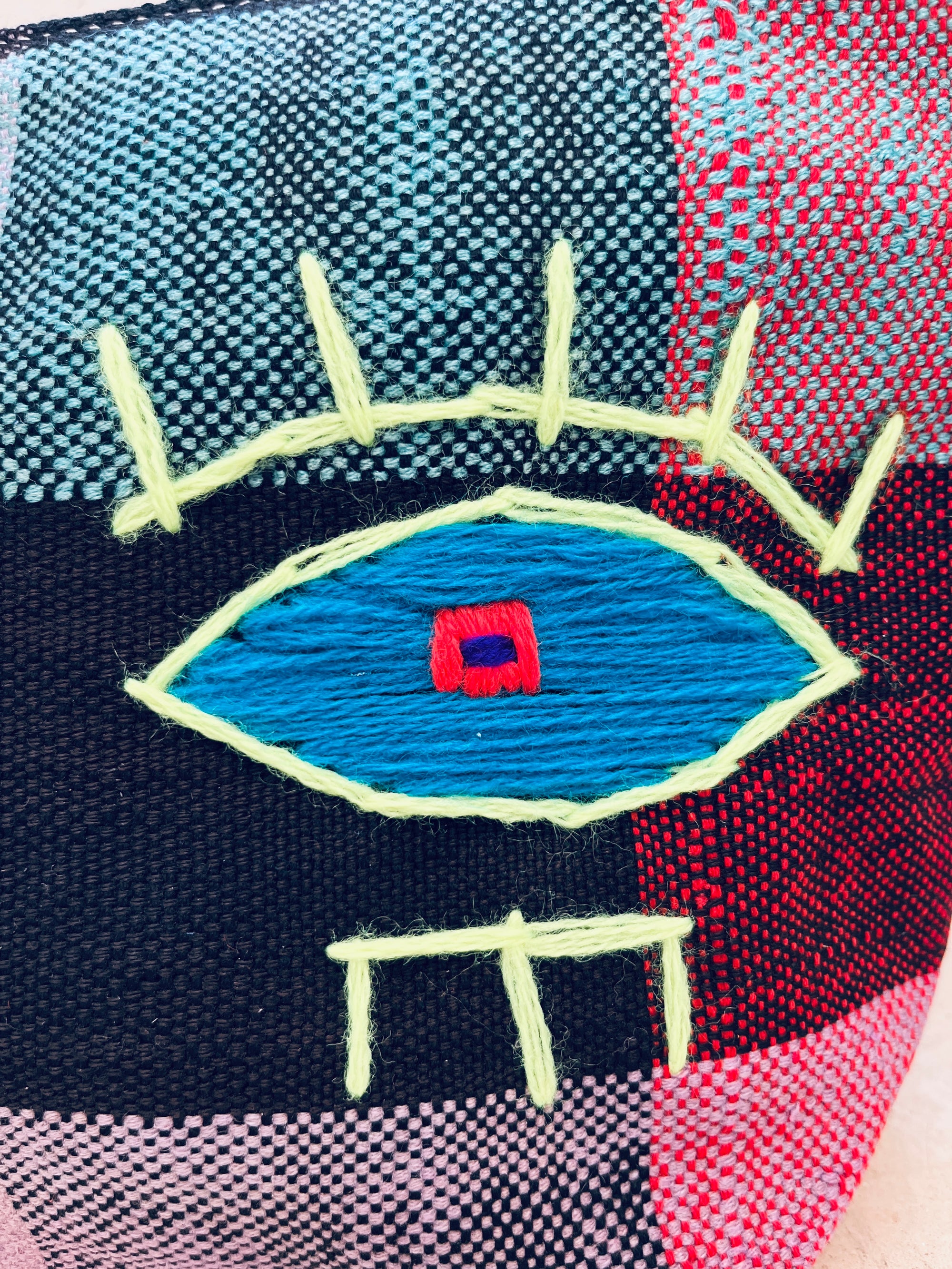 Eine Nahaufnahme der LEY! Tasche - Kosmetiktasche zeigt ein handgewebtes Patchwork mit einem gestickten blauen Augenmotiv, einer roten und marineblauen Mitte, gelben Wimpern und drei gelben Linien unterhalb des Auges, die an Zähne erinnern.