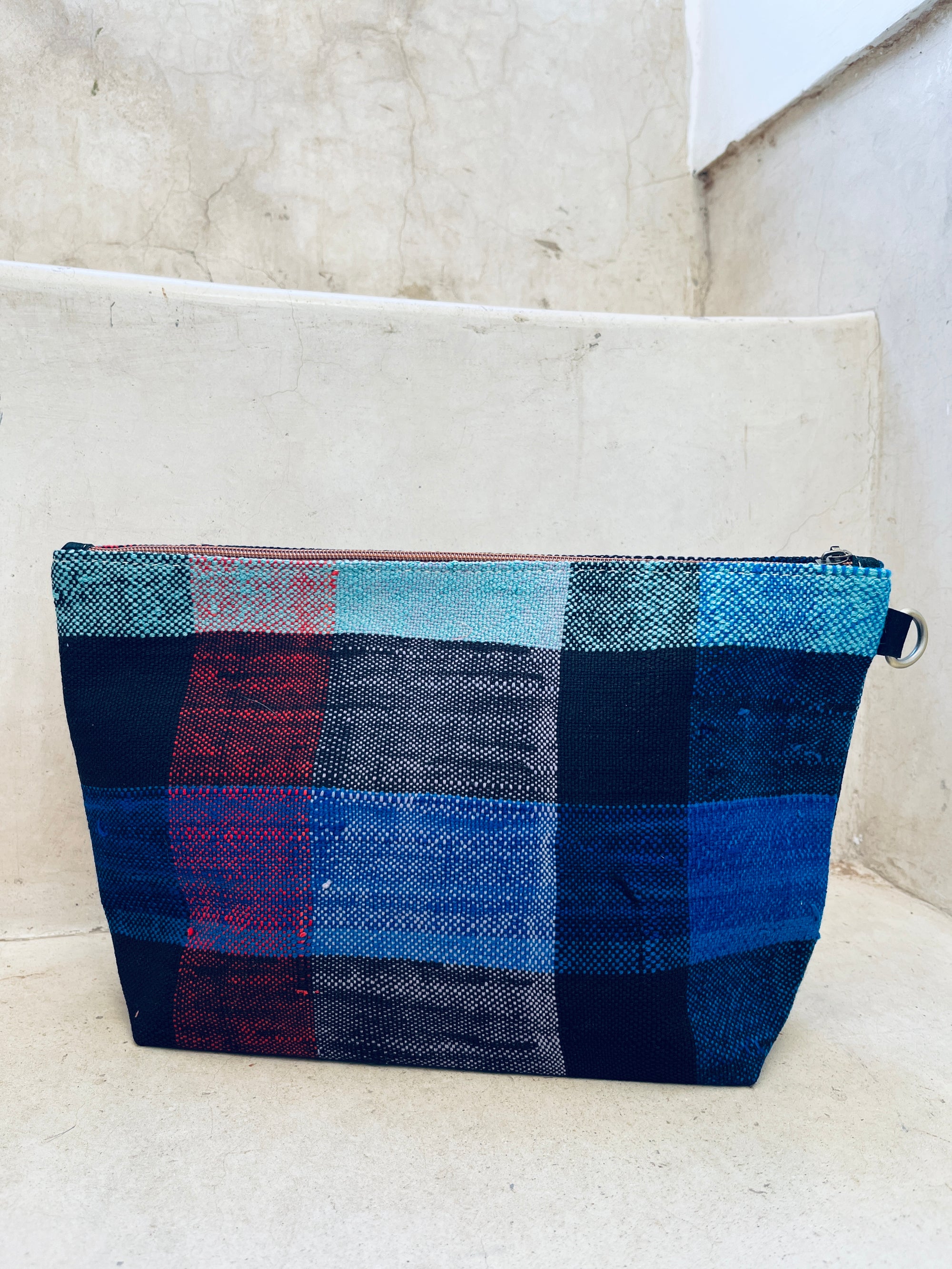 Die Tasche - Kosmetiktasche von LEY! ist handgewebt, rechteckig und zeigt ein farbenfrohes Plaid-Muster in Blau, Schwarz, Rot und Türkis. Sie liegt hier auf einer hellen Betontreppe.
