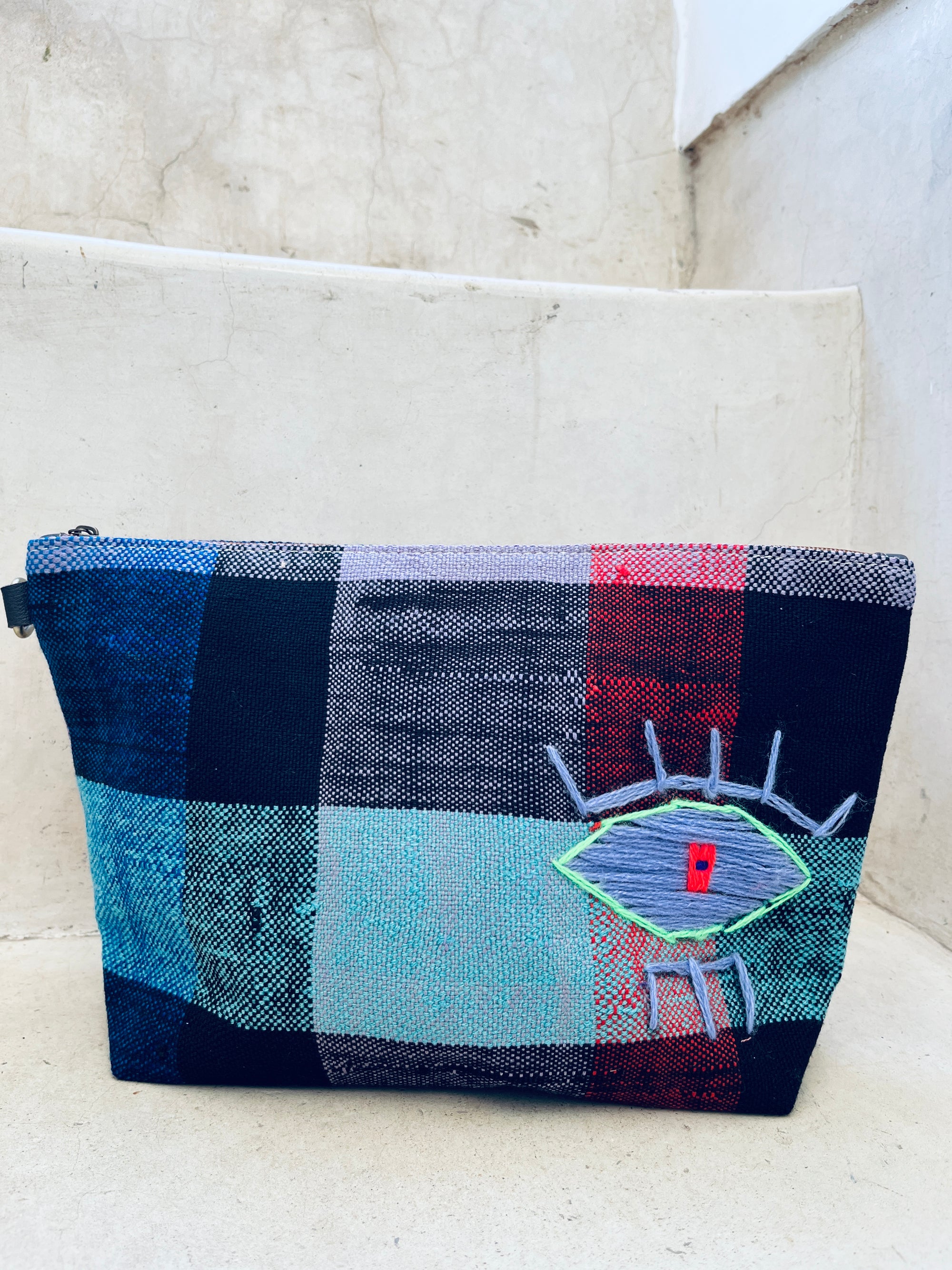 Die Kosmetiktasche "Tasche - Kosmetiktasche" von LEY! mit blau, schwarz, rot und türkis kariertem Muster sowie gesticktem Auge schmückt die hellen Betontreppen.