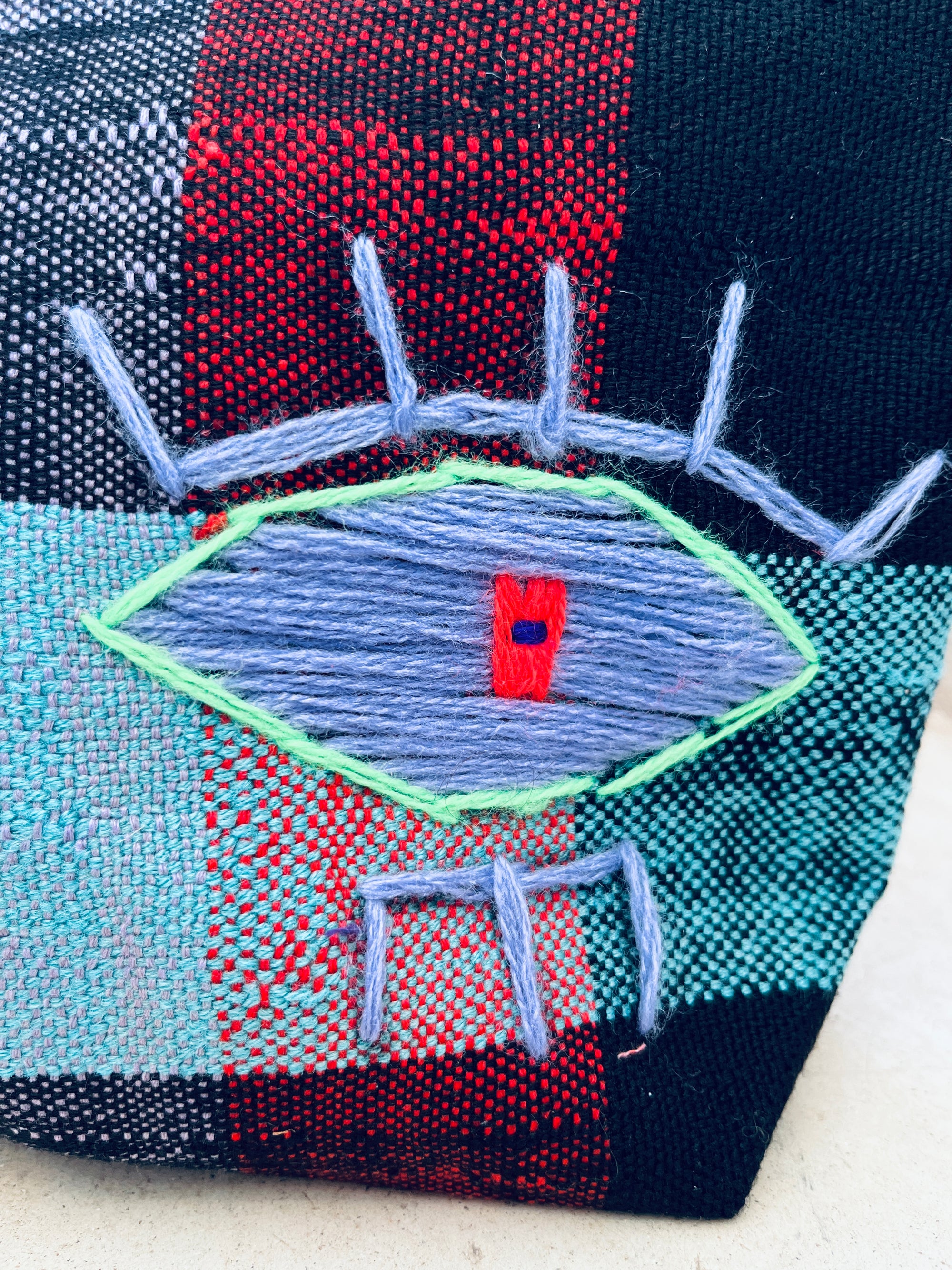 Eine Nahaufnahme eines gestickten Auges mit blauer Iris, roter quadratischer Pupille, grünem Umriss und blauen Wimpern auf kariertem Stoff, abgebildet auf der LEY! Tasche - Kosmetiktasche mit schwarzem, rotem und blaugrünem Hintergrund.