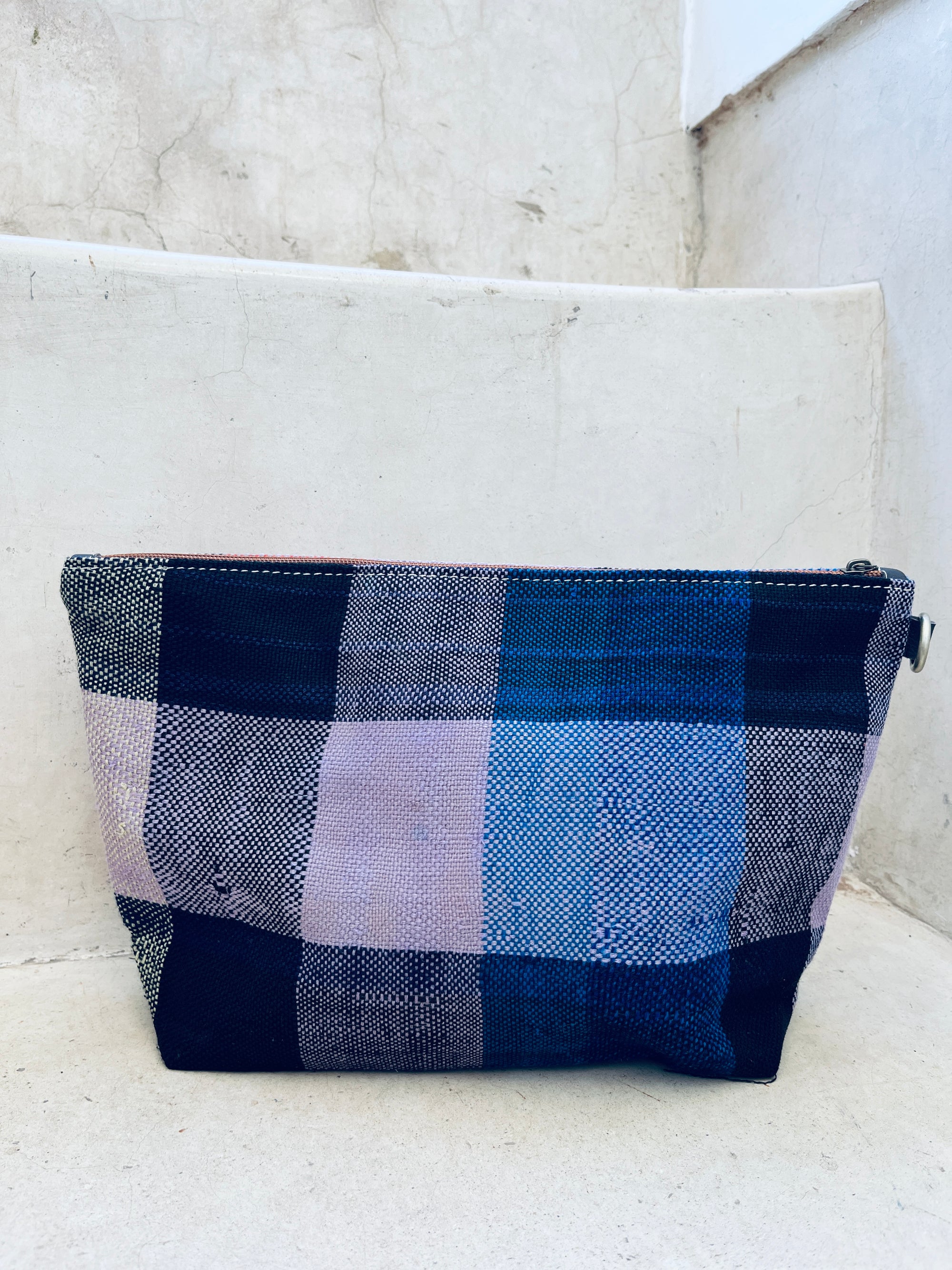 Die rechteckige Kosmetiktasche "Tasche - Kosmetiktasche" von LEY! mit blau-lila Karomuster steht auf hellen Betonstufen. Sie besitzt einen Reißverschluss und eine flache Basis für stabilen Stand.