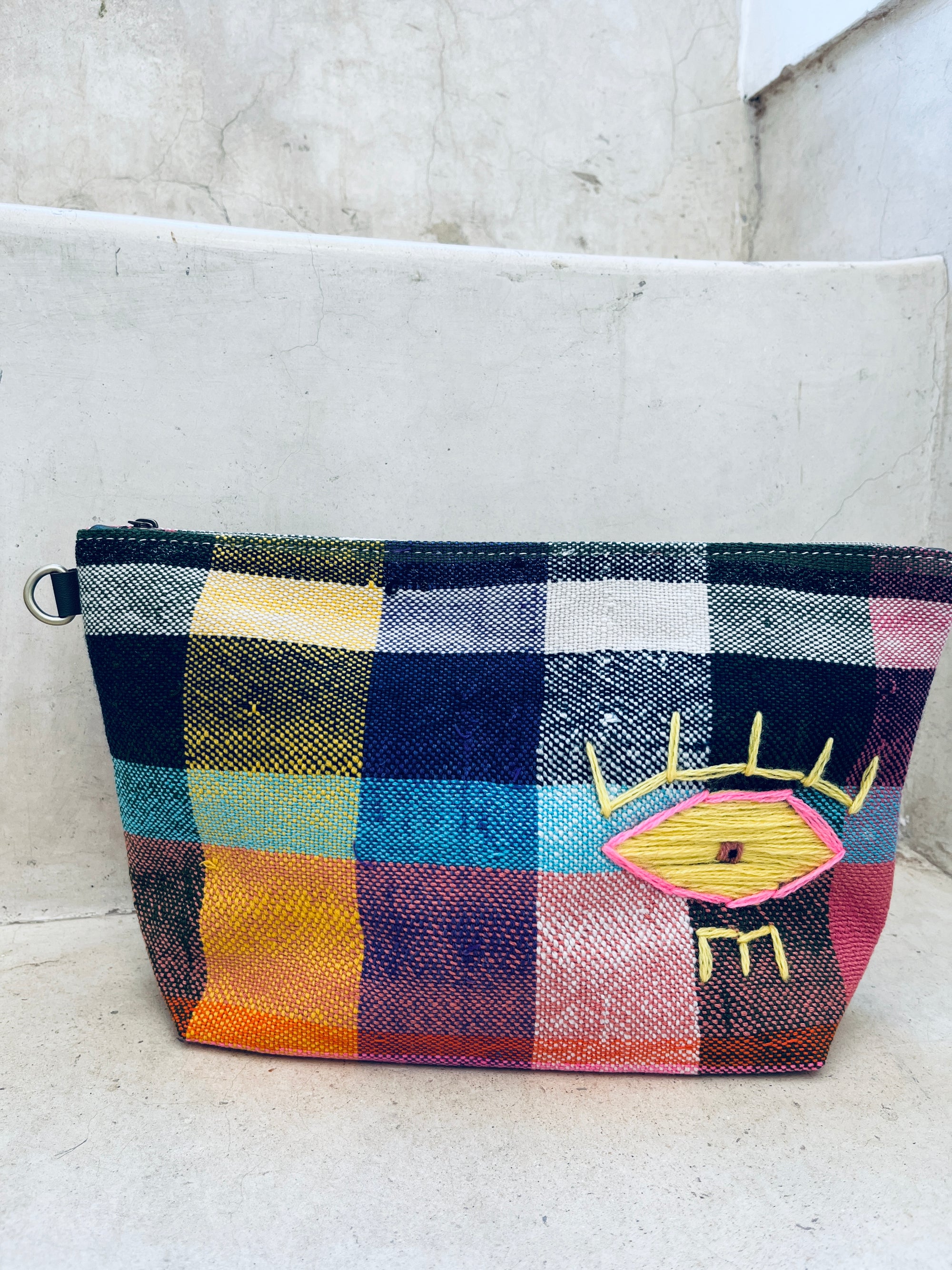 Eine leuchtend karierte Tasche - Kosmetiktasche von LEY! aus Marokko mit einem gelben und rosa Augenmotiv auf der Vorderseite, die auf einer hellen Betonstufe steht.