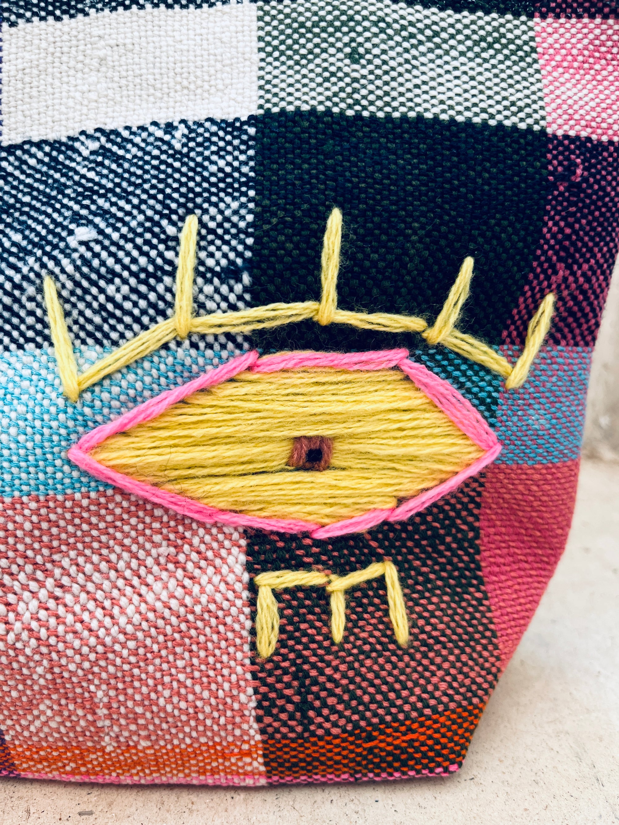 Eine Nahaufnahme der LEY! Tasche - Kosmetiktasche zeigt handgewebten, mehrfarbigen Stoff mit einem gelben, rosa und braunen gestickten Augenmotiv mit gelben Wimpern auf einem strukturierten, karierten Hintergrund, der von Marokko inspiriert ist.