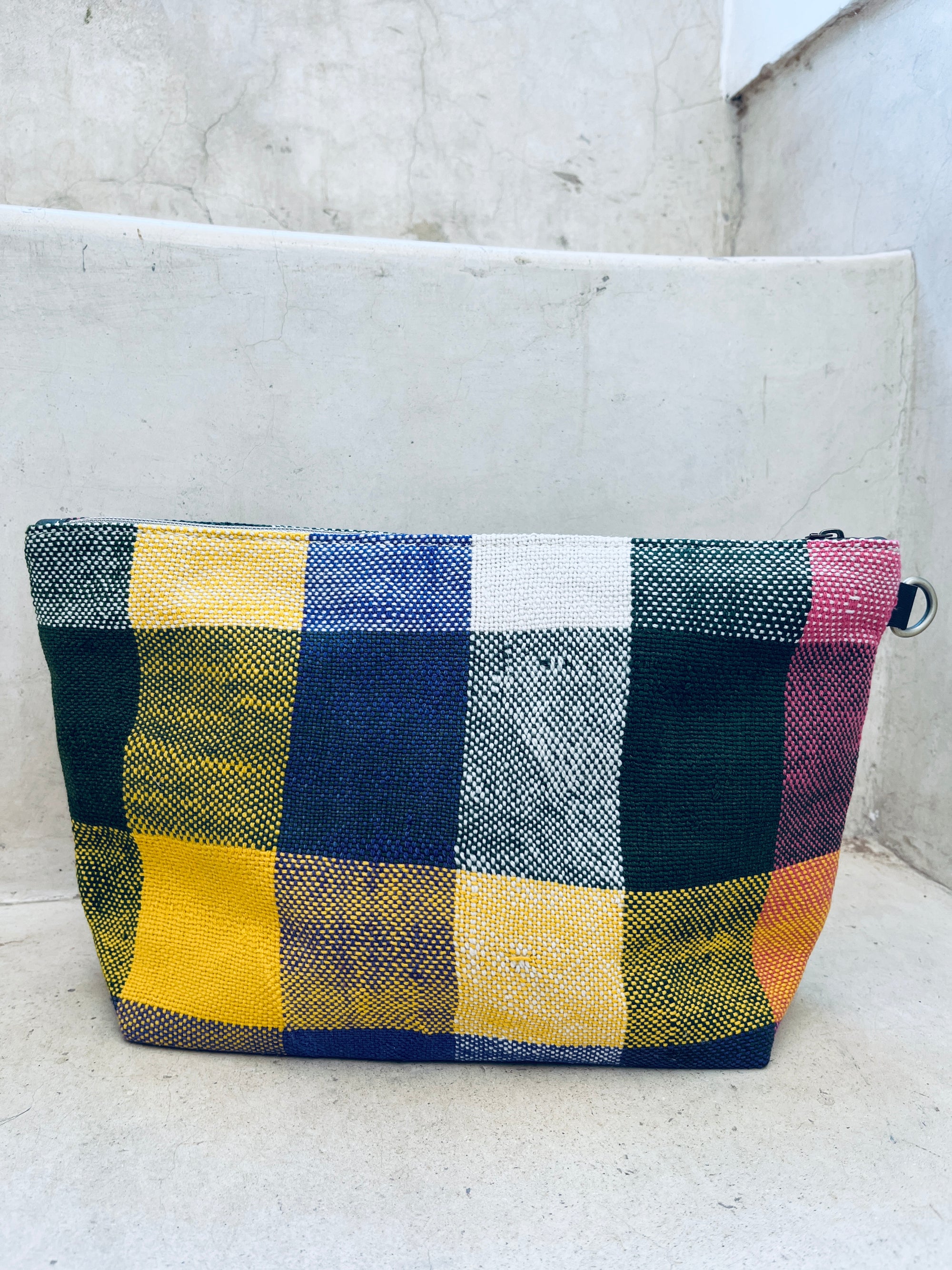 Die bunte, rechteckige Kosmetiktasche "Tasche - Kosmetiktasche" von LEY! mit großem handgewebtem Karomuster in Gelb, Blau, Grün, Weiß und Pink aus Marokko liegt auf weißen Betontreppen.