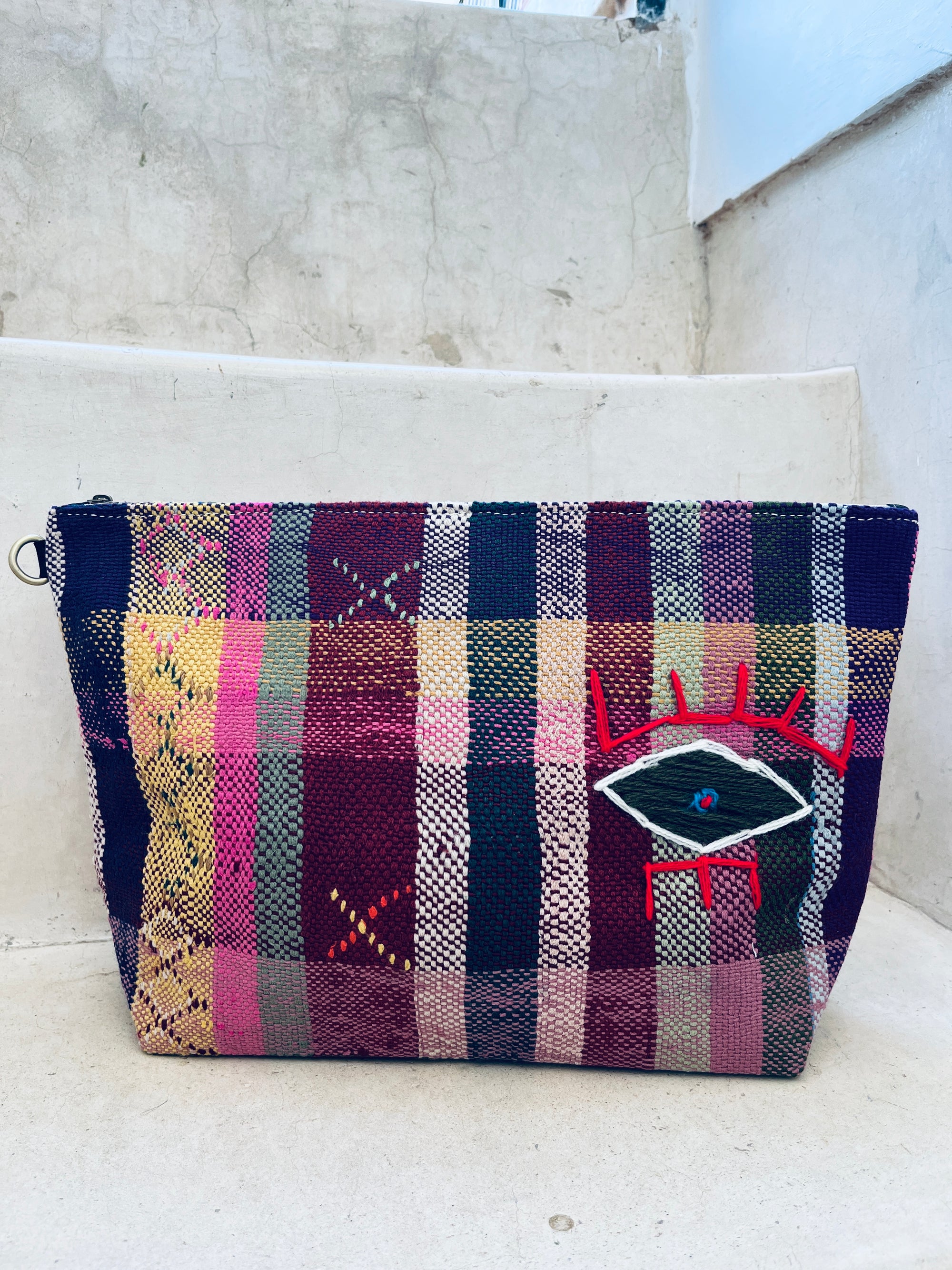 Eine leuchtende, handgewebte LEY! Tasche - Kosmetiktasche mit einem lila, rosa, gelben und grünen Karomuster ruht auf Betonstufen. Die Vorderseite zeigt ein gesticktes Auge mit roten Wimpern und einem blauen Punkt.