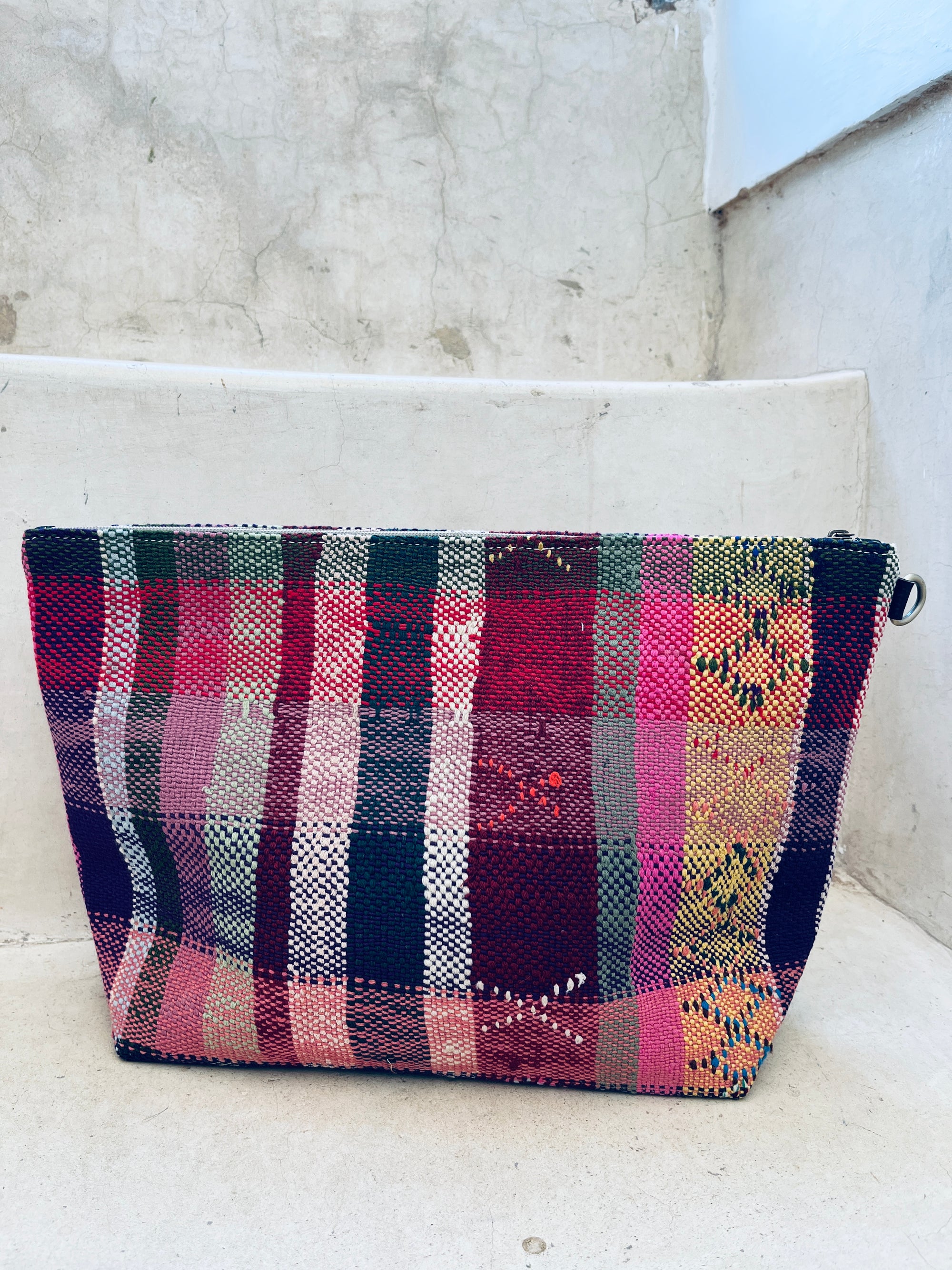 Die LEY! Kosmetiktasche besticht im bunten Karomuster mit Rot, Grün, Pink, Gelb und Navy auf leicht strukturiertem Stoff und sitzt auf weißen Betonstufen.