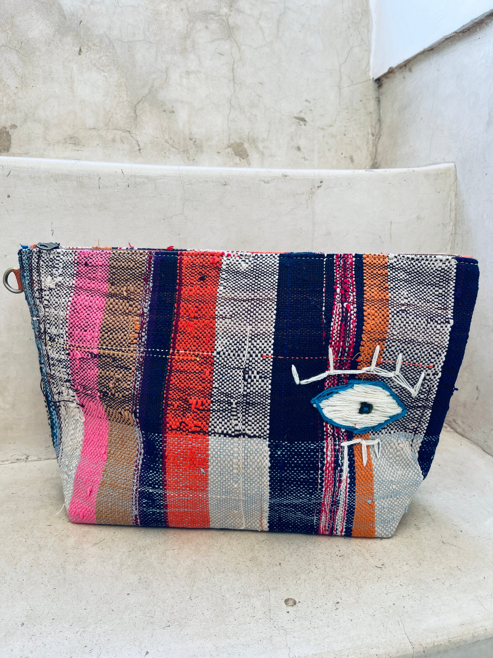 Die LEY! Tasche - Kosmetiktasche ist eine farbenfrohe, handgewebte Tasche mit vertikalen rosa, roten, marineblauen, orangefarbenen und weißen Streifen und einem aufgestickten blauen und weißen Auge, die auf Steinstufen vor einer strukturierten Wand ausgestellt ist.
