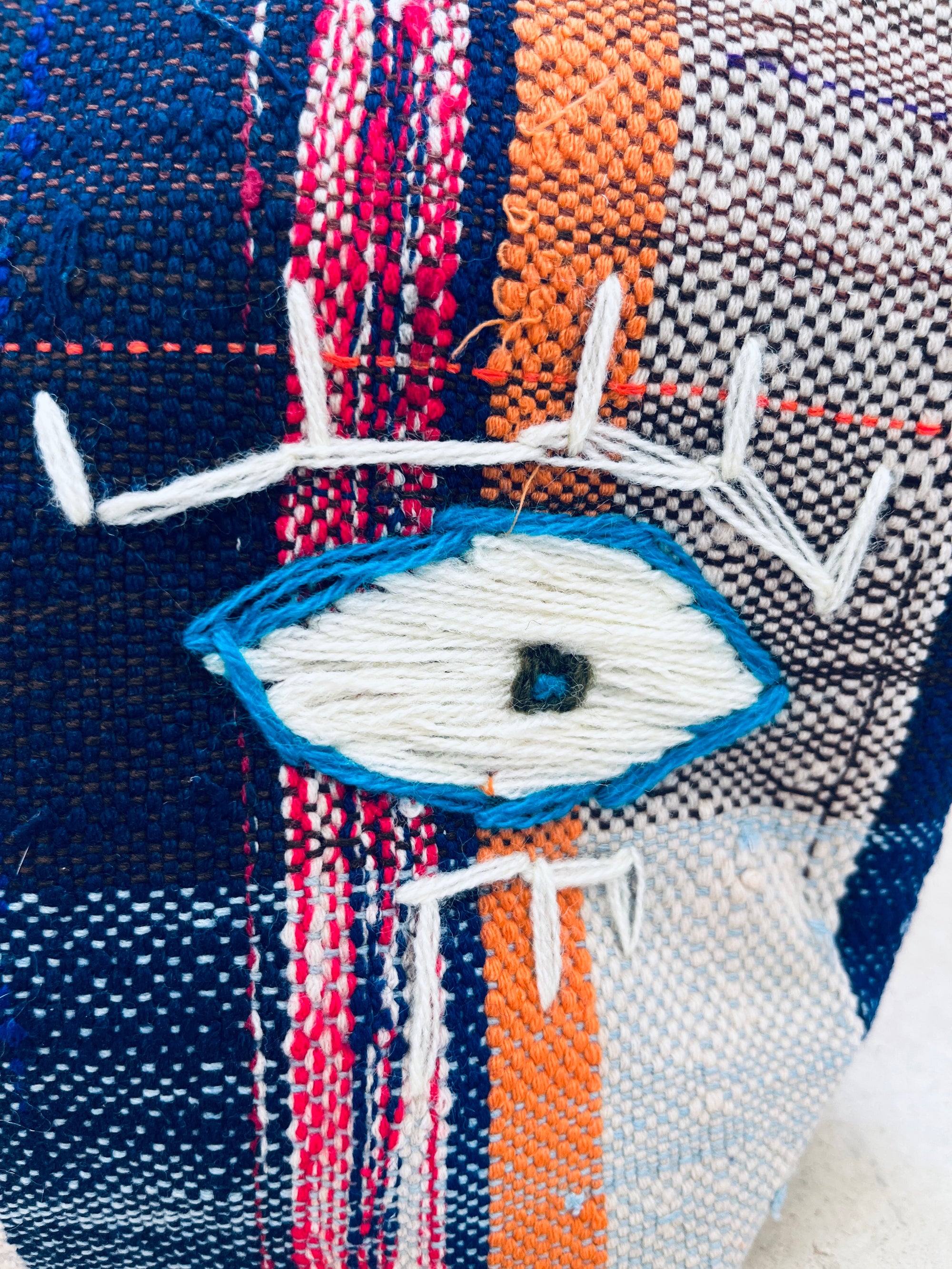 Eine Nahaufnahme eines handgewebten Textils mit einem aufgestickten blau-weißen Augenmuster, perfekt für die LEY! Tasche - Kosmetiktasche, mit orangefarbenen, marineblauen und weißen Mustern, die von Marokko inspiriert sind.