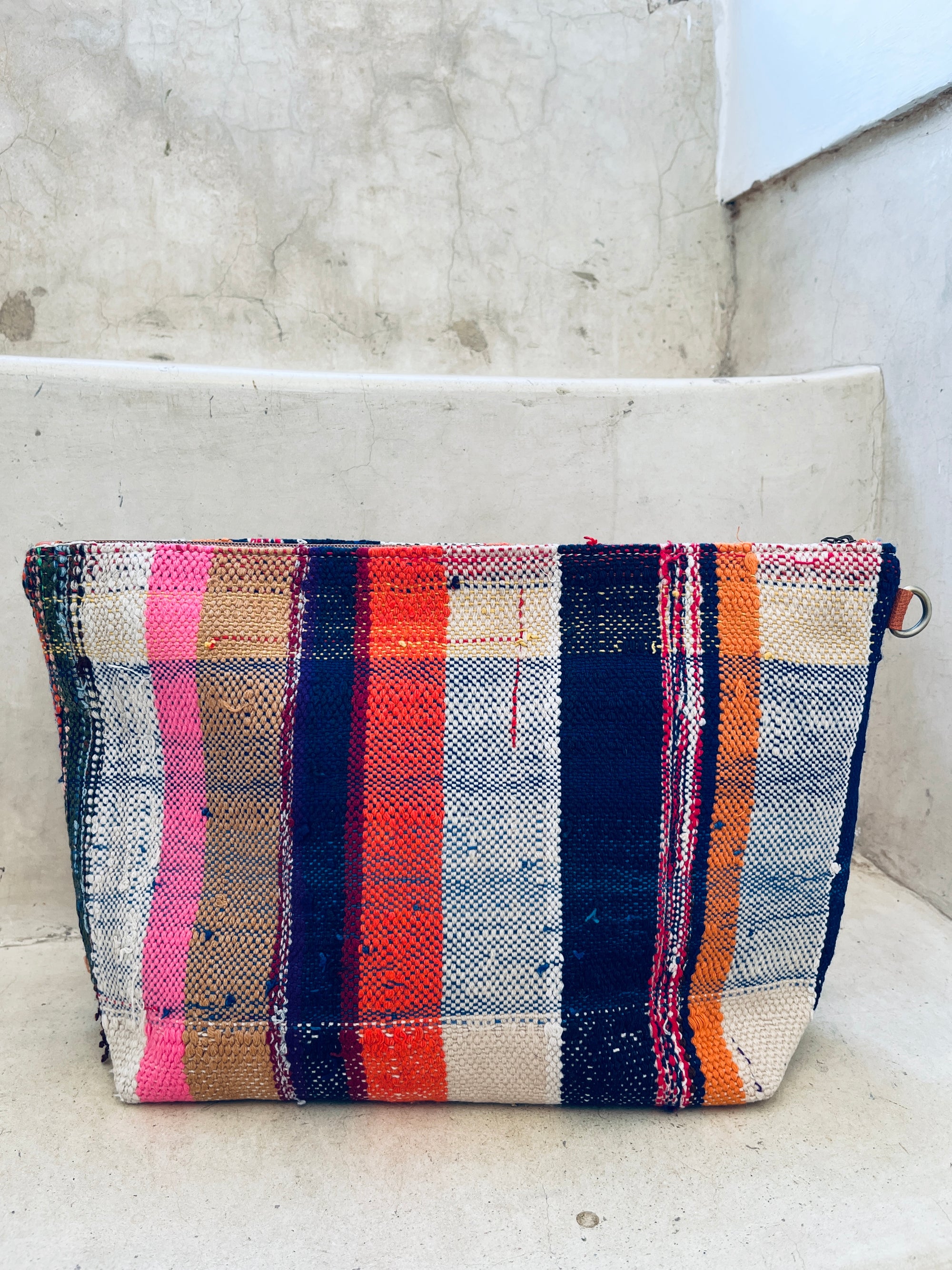 Die handgewebte Kosmetiktasche von LEY! mit vertikalen Streifen in Pink, Marineblau, Orange, Beige und Creme liegt auf hellen Steinstufen vor einer strukturierten Steinwand.