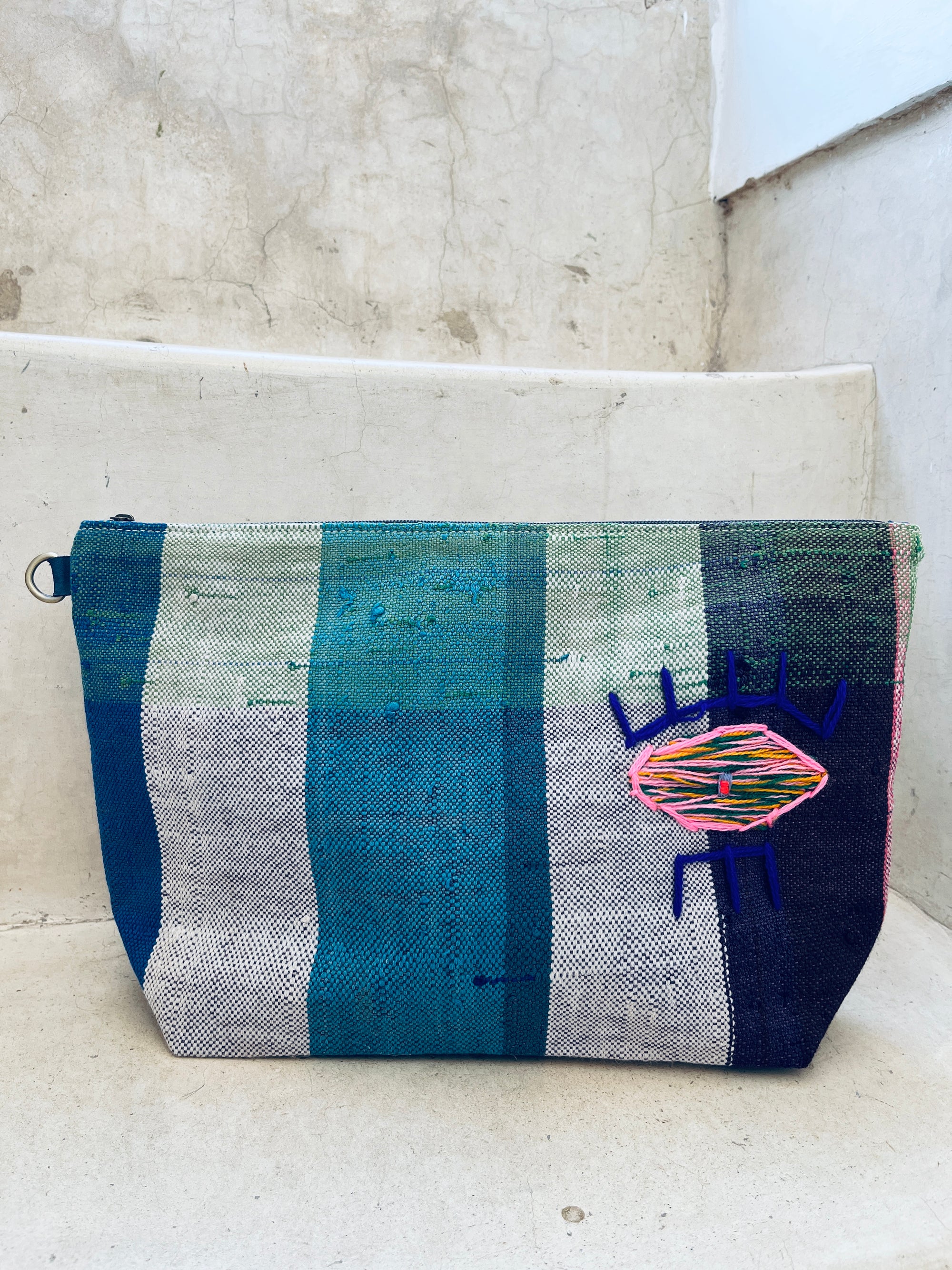 Die rechteckige Kosmetiktasche "Tasche - Kosmetiktasche" von LEY! aus Marokko besticht mit blauen, grünen und weißen Streifen sowie einem bunten gestickten Käfermotiv und liegt auf einer Betontreppe.