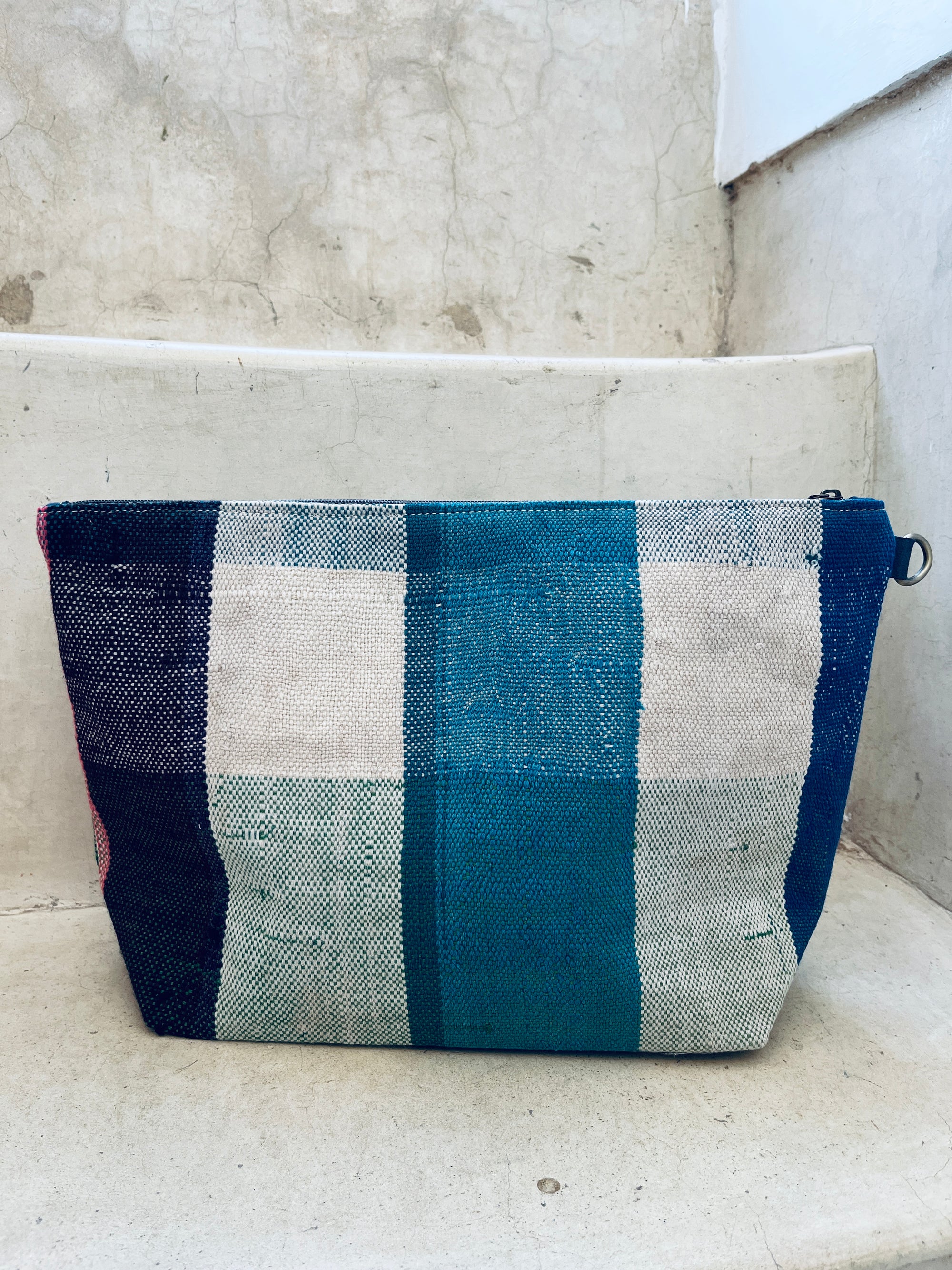 Die rechteckige, handgefertigte LEY! Kosmetiktasche mit gewebtem blau-türkis-weiß kariertem Muster steht auf einer Betontreppe vor einer strukturierten Wand.