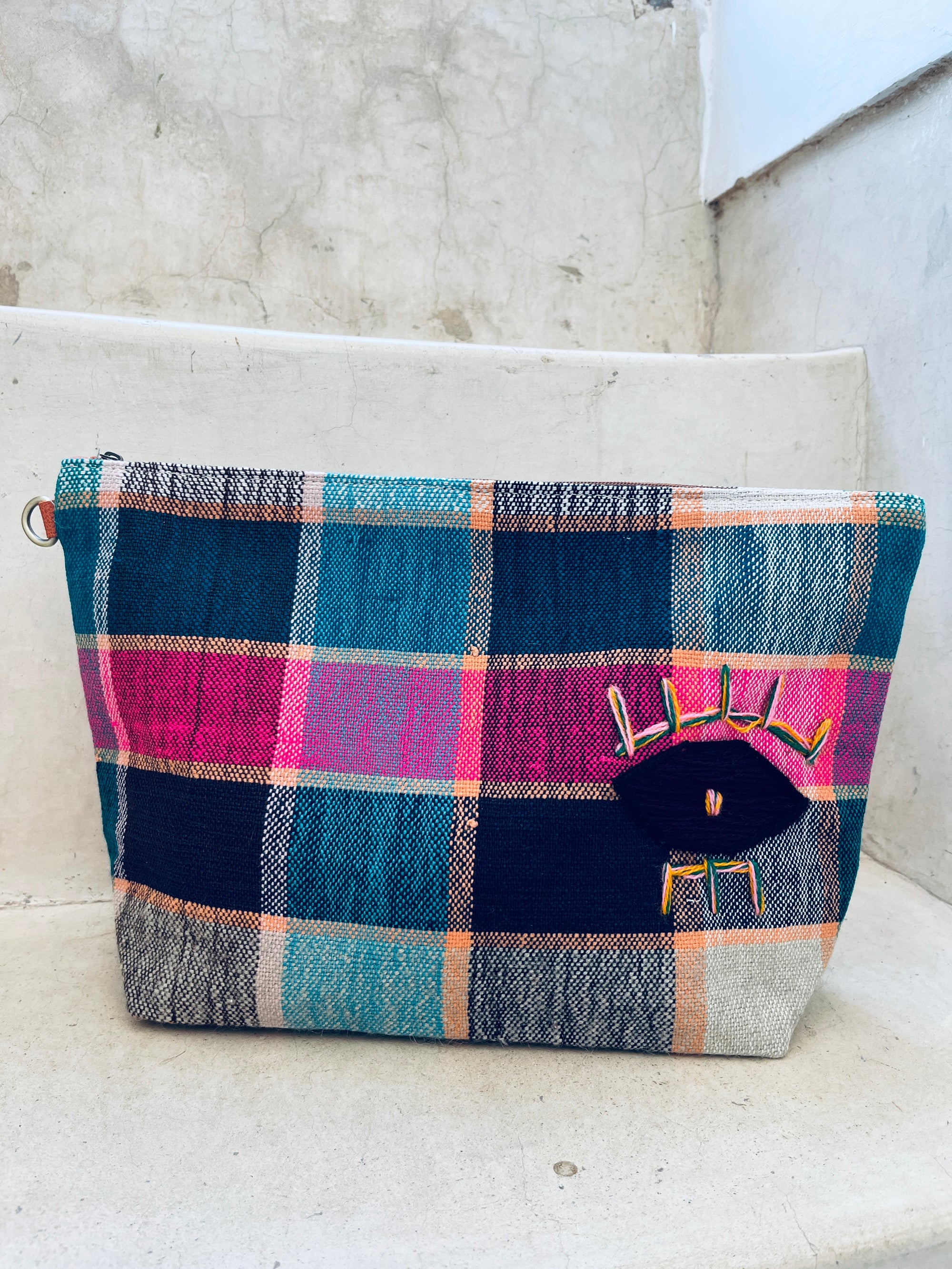 Die LEY! Tasche - Kosmetiktasche zeigt blaue, pinke, türkise und weiße Karos sowie ein auffälliges Augenmotiv in Gelb, Pink und Schwarz. Sie liegt auf einer off-whitefarbenen Betontreppe.