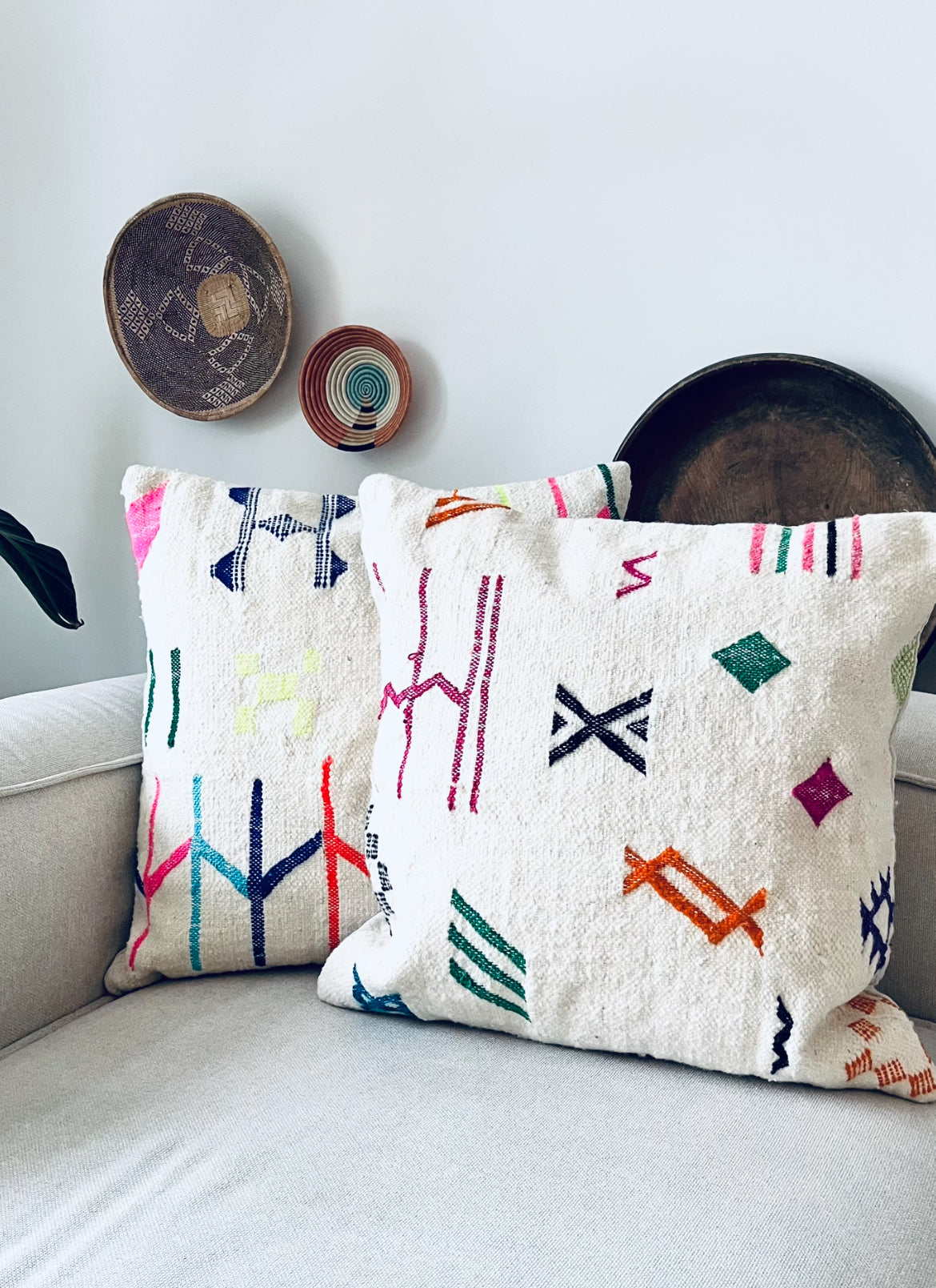 Zwei weiße Kissen mit bunten geometrischen Mustern liegen auf einem hellgrauen Sofa. Dekorative geflochtene Körbe hängen an der weißen Wand dahinter und verleihen dem Raum einen gemütlichen und künstlerischen Touch.
