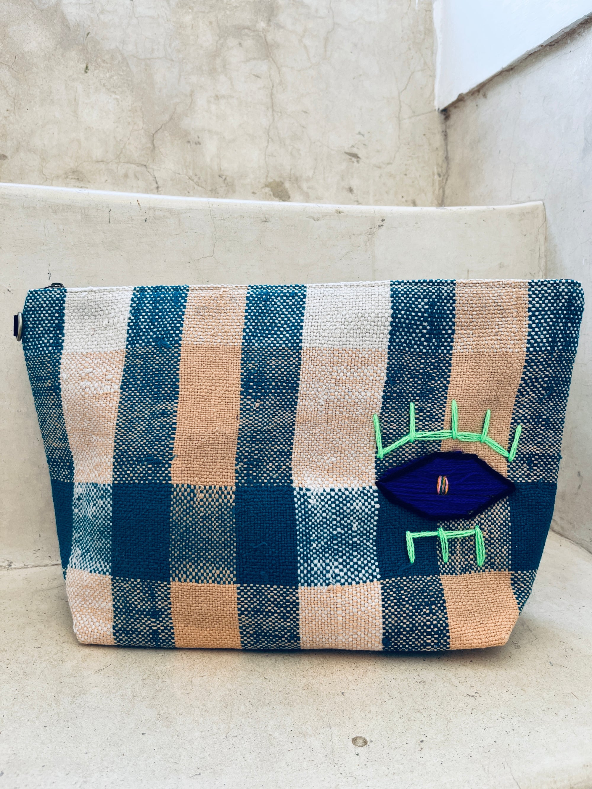 Die LEY! Tasche - Kosmetiktasche mit blau-beigem Karomuster, auffälligem Auge-Patch und neon-grüner Stickerei liegt auf einer Betontreppe.