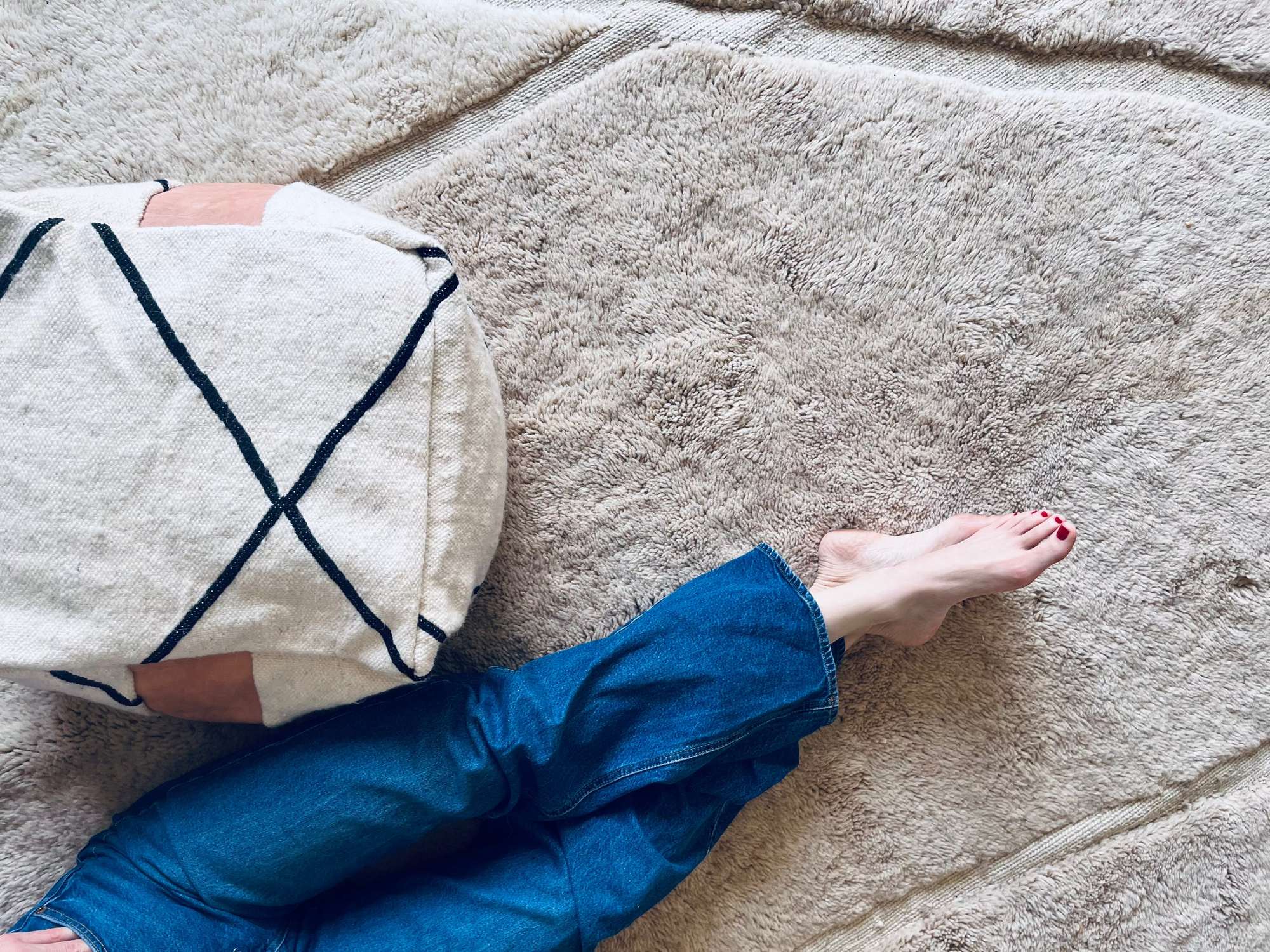 Eine Person in blauen Jeans sitzt mit nackten Füßen auf einem hellen strukturierten Teppich neben einem weißen Kissen mit schwarzen geometrischen Linien. Nur die Beine und der untere Teil des Körpers sind sichtbar.