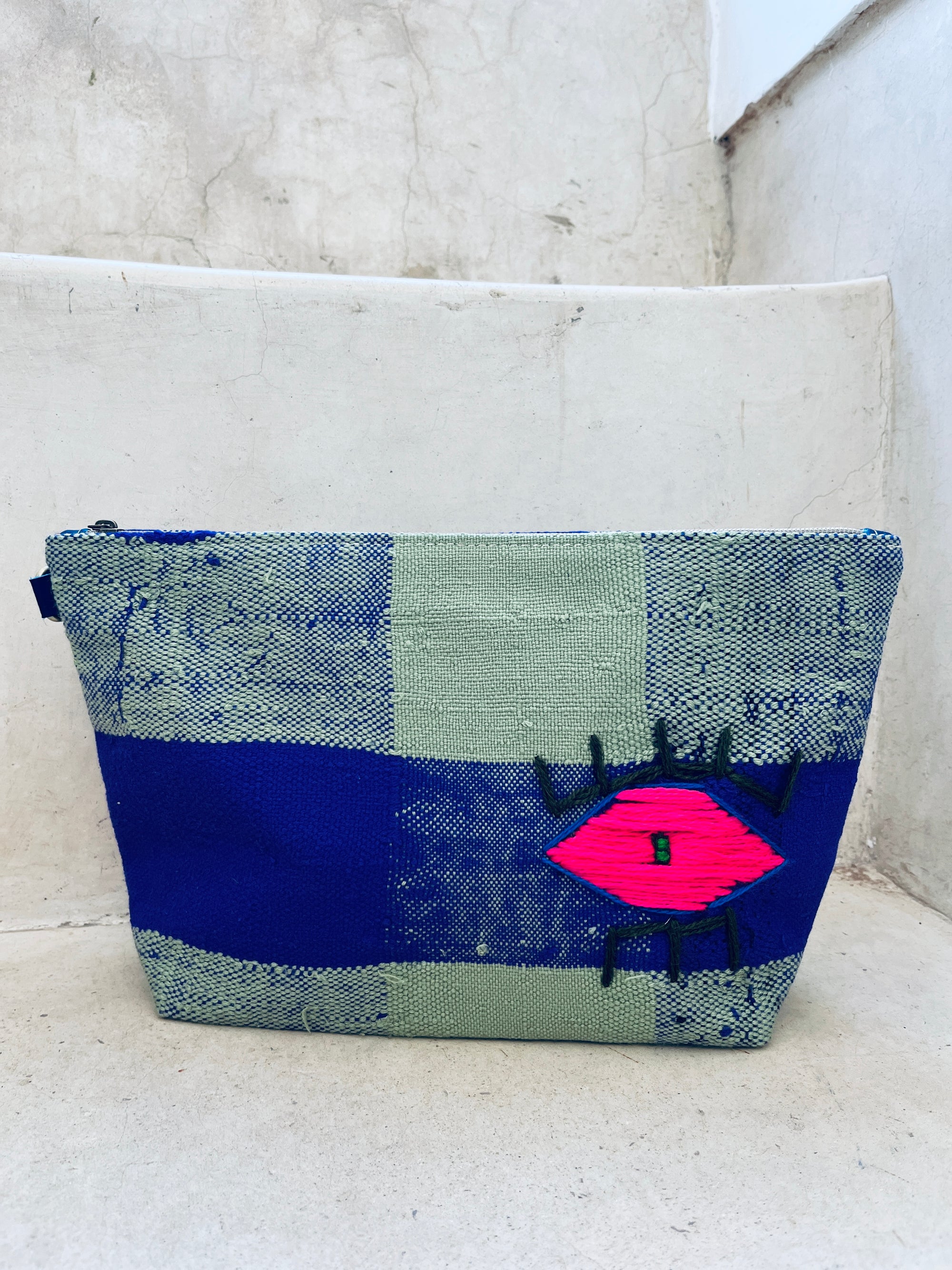 Die Tasche - Kosmetiktasche von LEY! ist handgewebt in Marokko, zeigt ein blau-graues Karomuster und ein pinkes Auge-Motiv, und liegt auf einer hellen Steinstufe.
