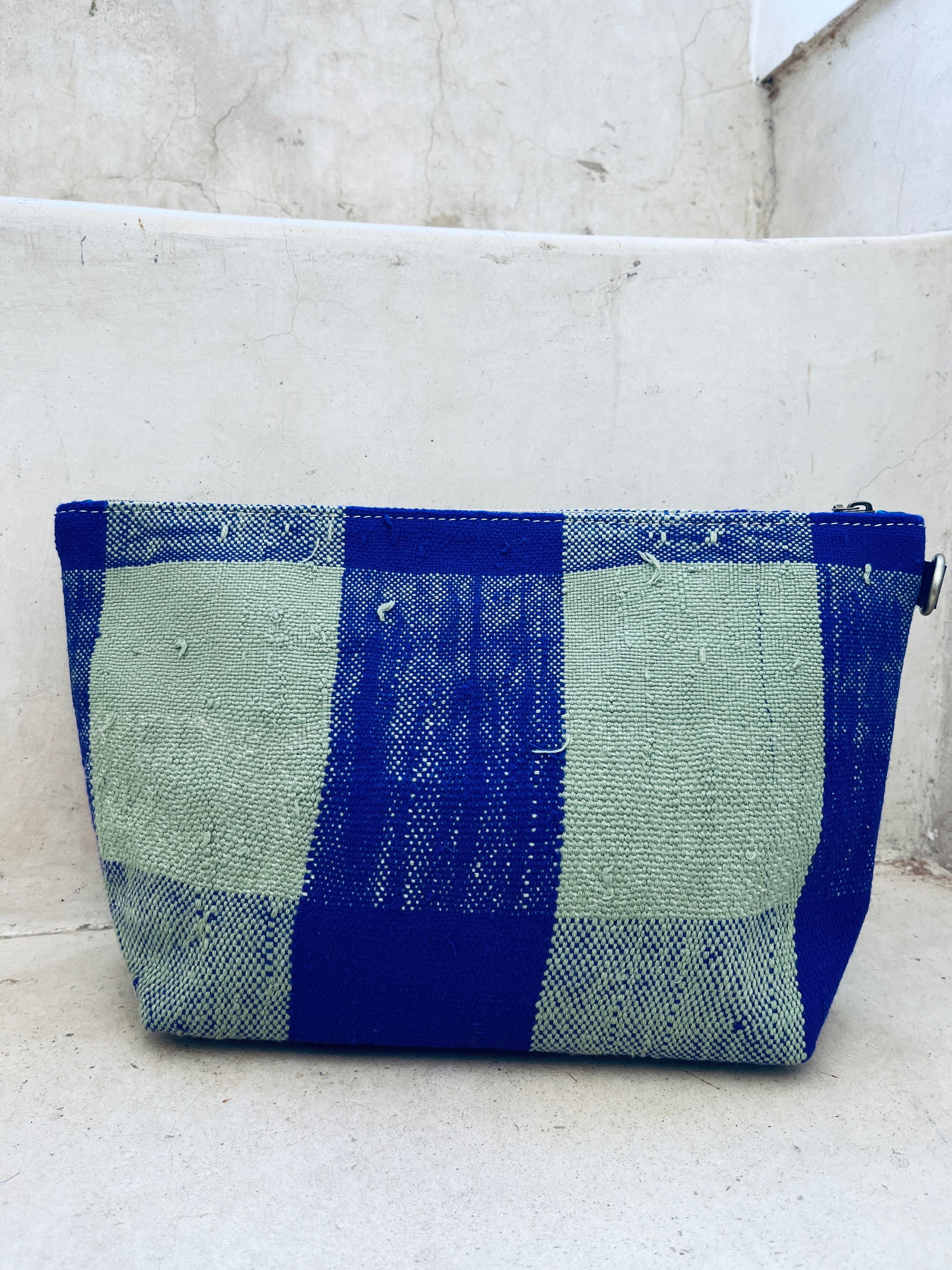 Die rechteckige, handgefertigte Tasche - Kosmetiktasche von LEY! mit Reißverschluss im blau-grünen Karo liegt auf weißen, rissigen Betontreppen.
