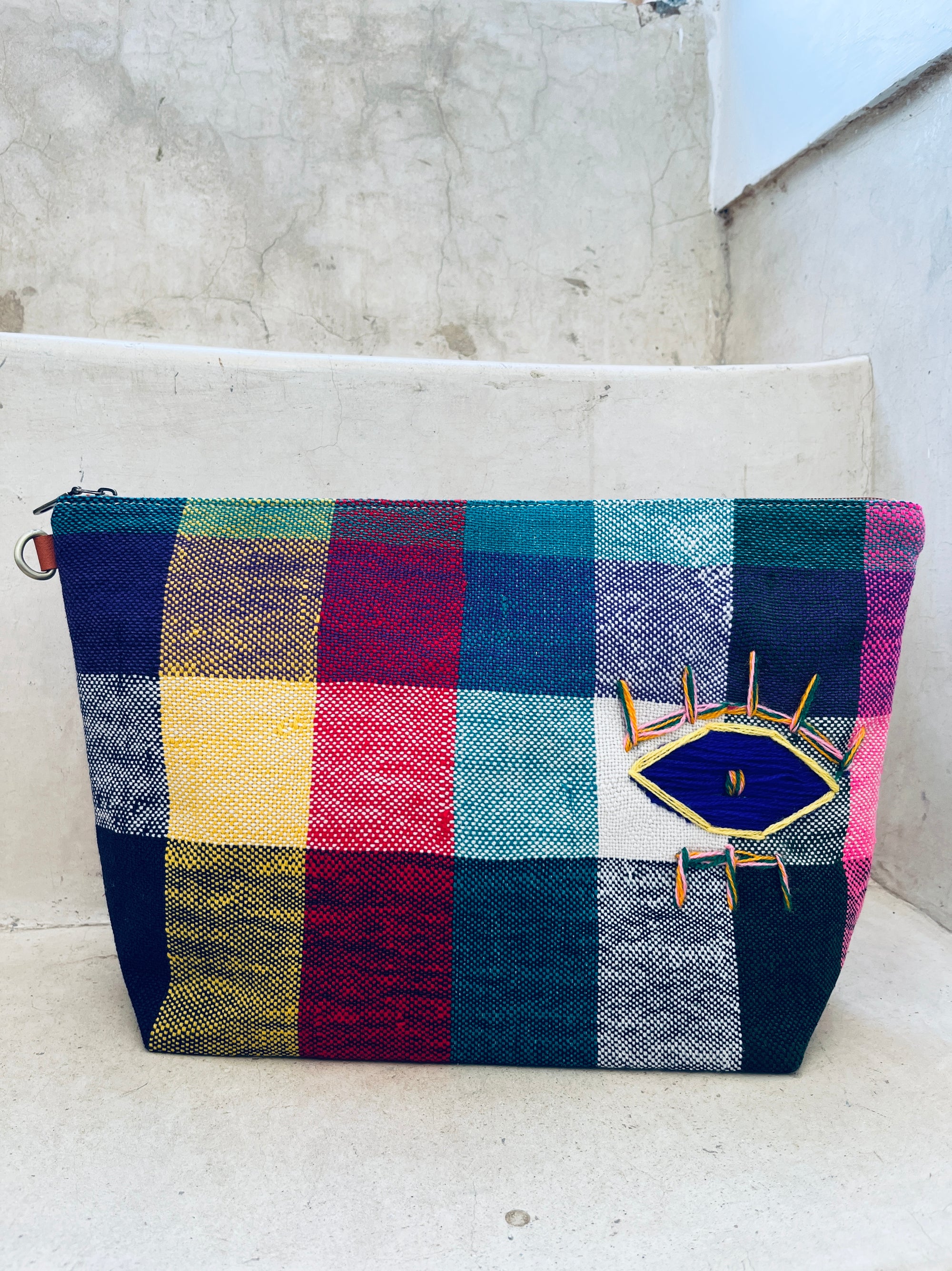 Eine farbenfrohe Tasche - Kosmetiktasche von LEY! aus dem Hause Marokko sitzt auf einer weißen Treppe und zeigt ein auffälliges Augenmotiv mit gelber, blauer und rosa Stickerei auf der Vorderseite.