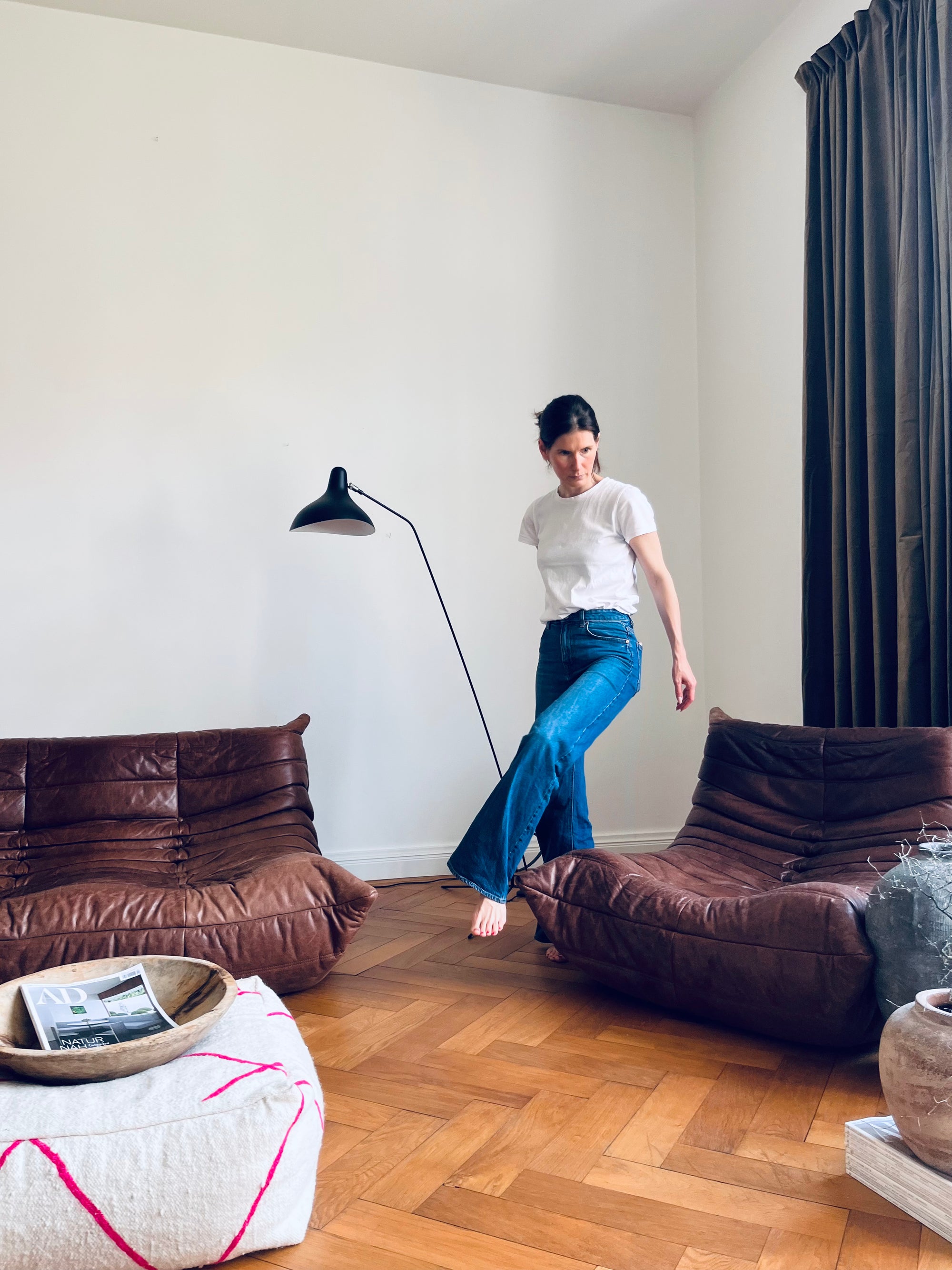 Eine Frau in weißem T-Shirt und blauen Jeans steht barfuß auf der Kante eines braunen Ledersofas in einem modernen Wohnzimmer mit Holzfußboden, einer Stehlampe und neutraler Einrichtung.
