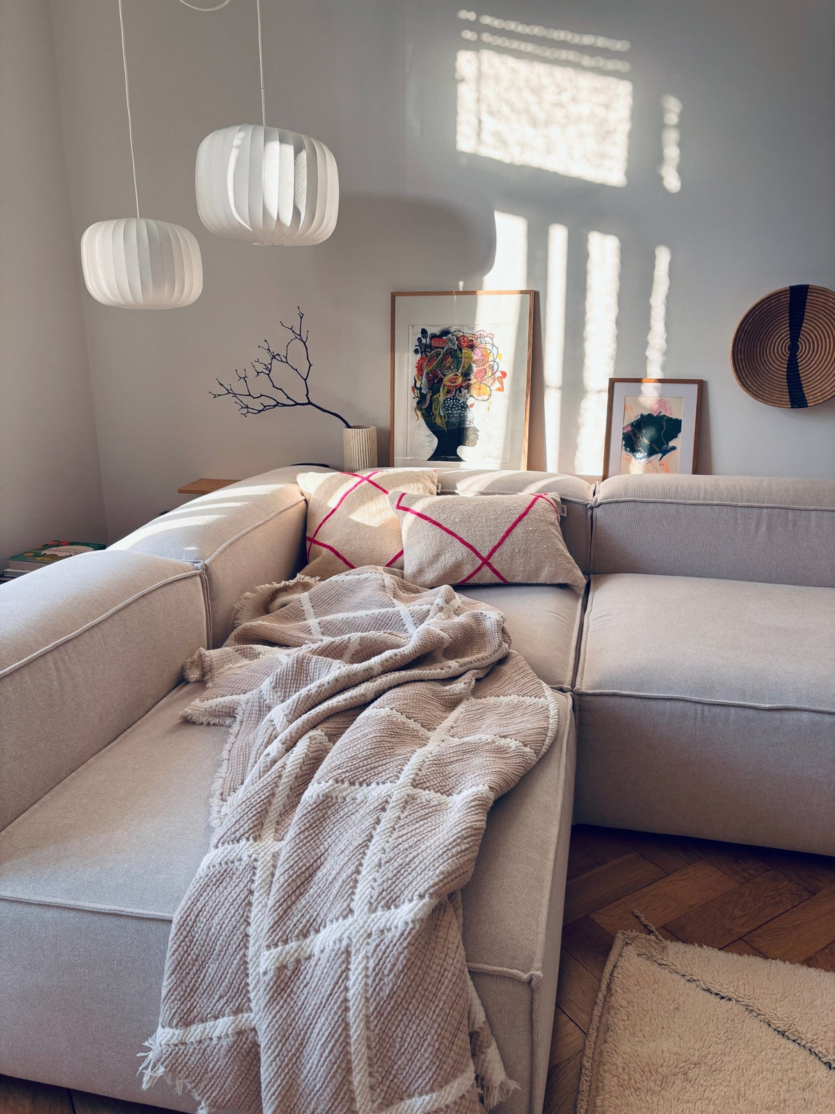 Beiges Ecksofa mit LEY!-Kissen – Classic Line Mina 30x50, eine Decke, Wanddekoration und hereinströmendes Sonnenlicht.