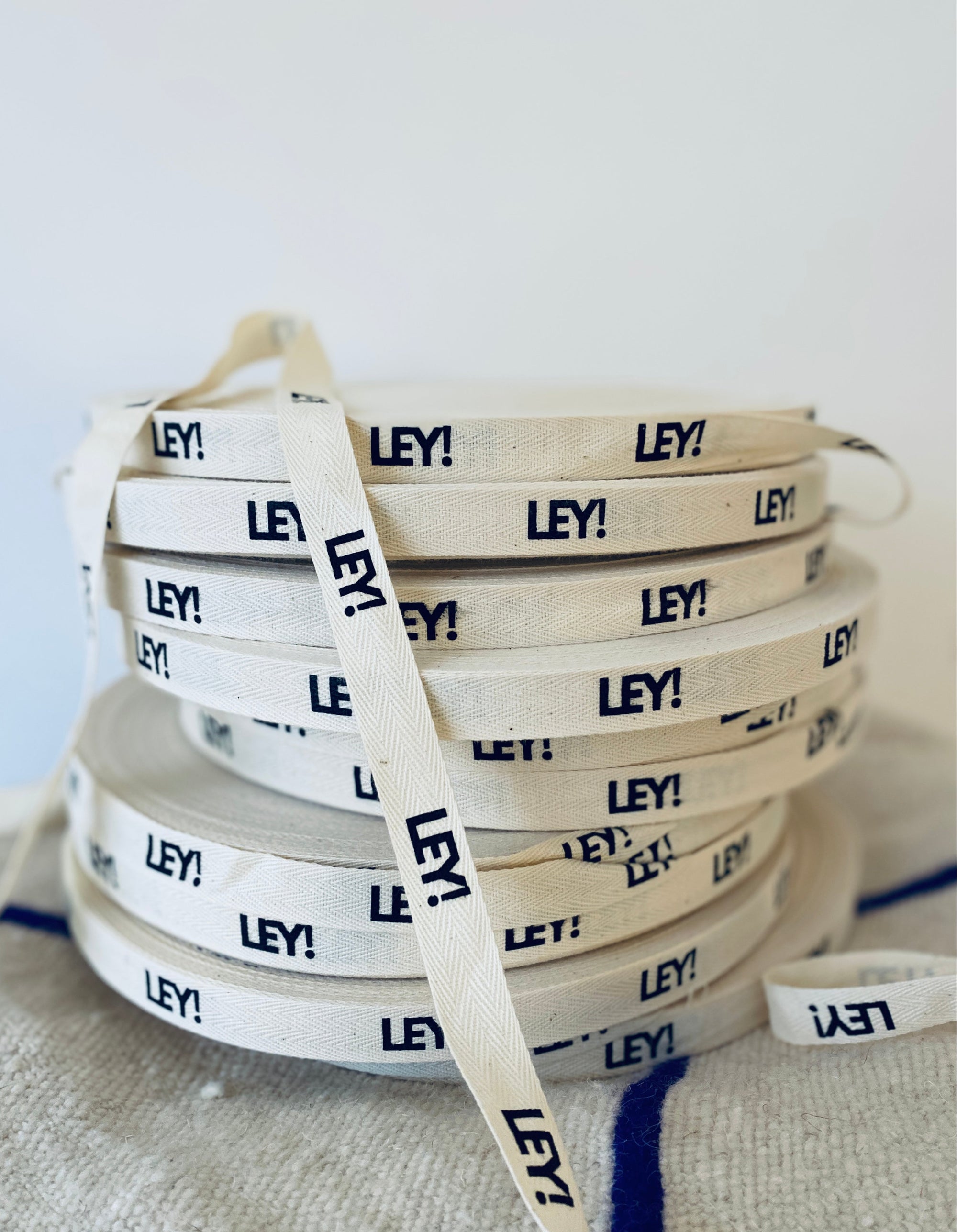 Ein Stapel beigefarbener Stoffbänder, die wiederholt mit dem Wort "LEY!" in fetten schwarzen Buchstaben bedruckt sind, vor einem hellen, neutralen Hintergrund.