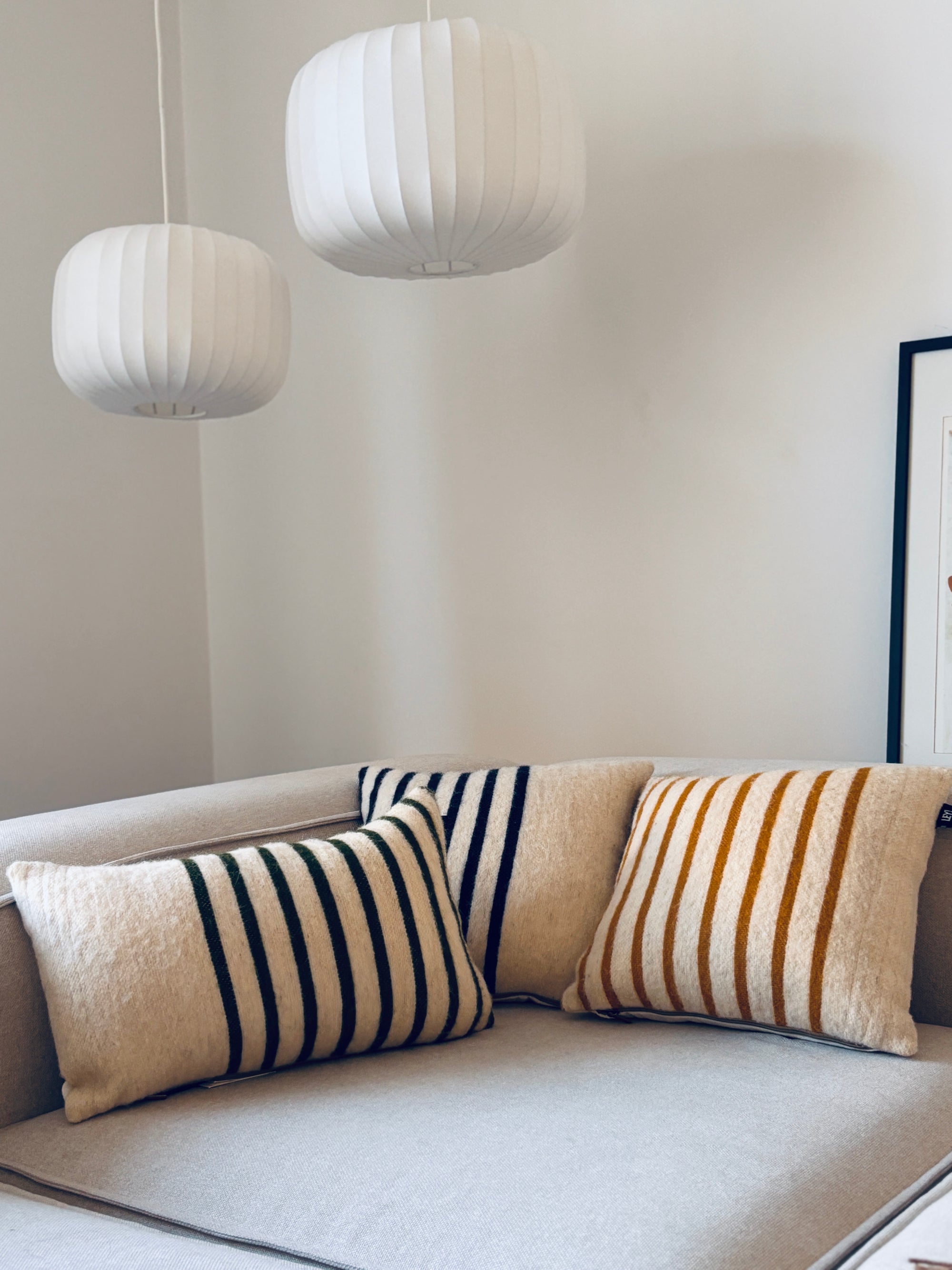 Ein hellgraues Sofa mit zwei gestreiften Kissen - eines grün, eines LEY! Kissen WINTER EDITION TANNE 30 x 50 in Gelb - steht unter drei modernen weißen Pendelleuchten vor einer schlichten weißen Wand.