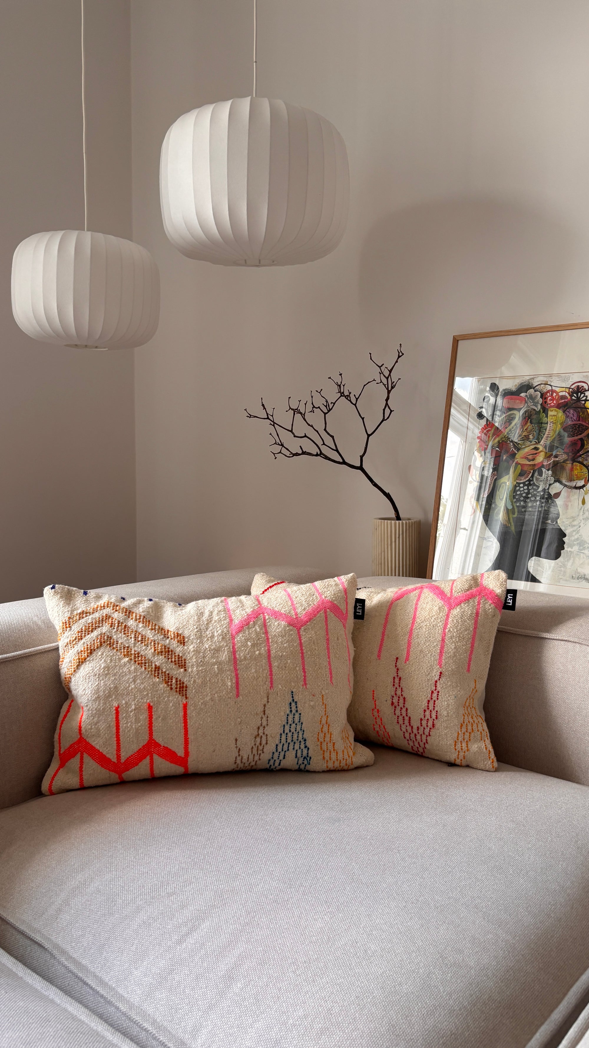 Ein beiges Sofa mit zwei LEY! Kissen Colorful Line, Hängelampen, gerahmter Kunst und einem kahlen Zweig in einer Vase.